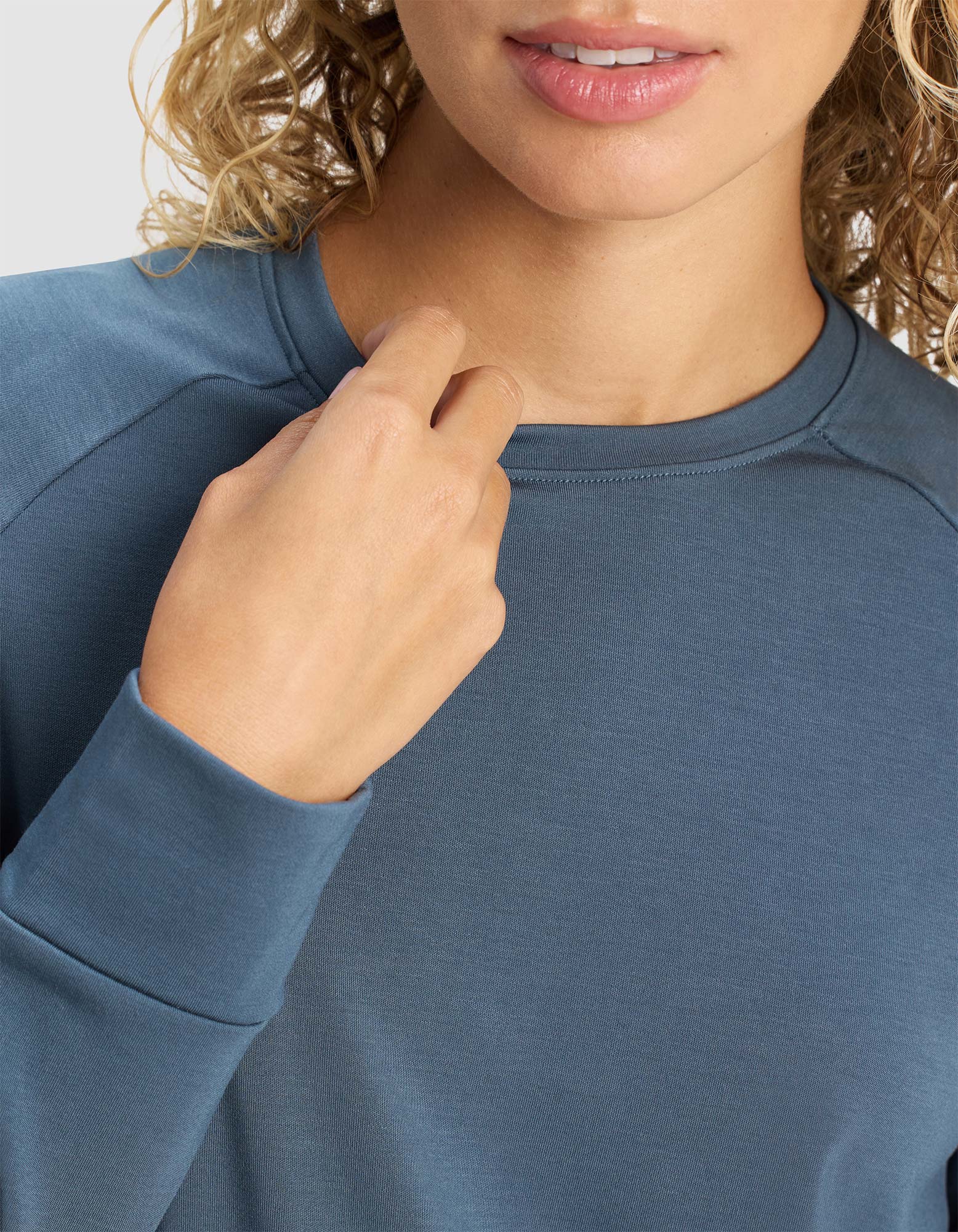 A woman with curly blonde hair wears a blue Cozy Earth Women's Brushed Bamboo Pullover Crew, holding the collar with one hand; only her shoulders to lips are visible.
