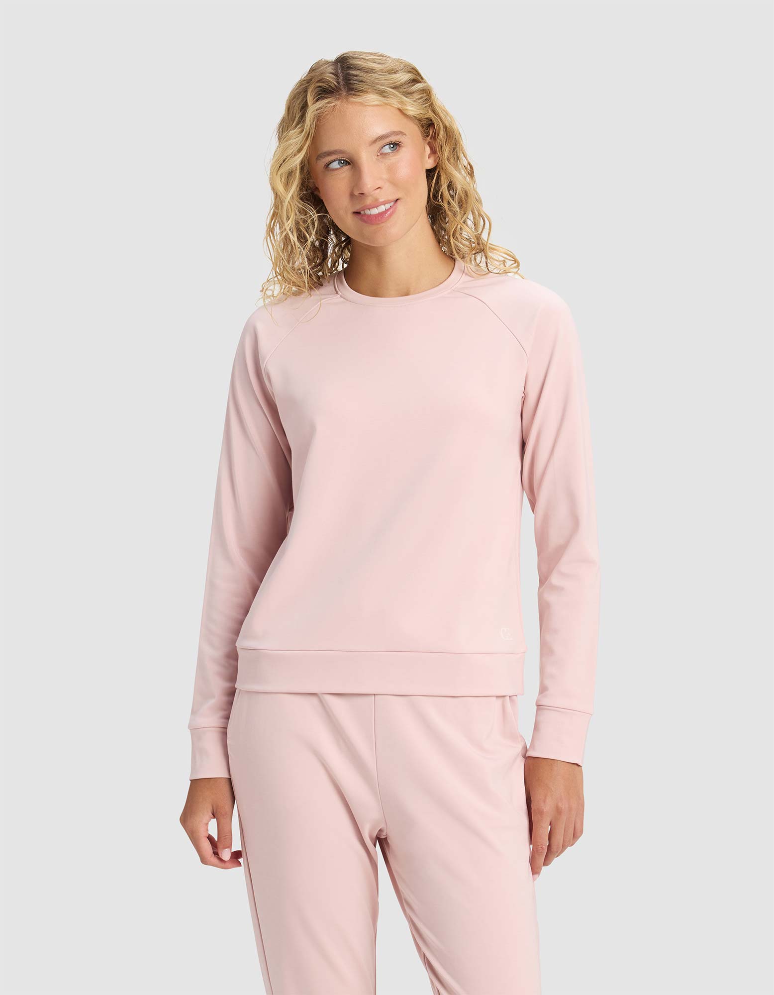A woman with blonde, wavy hair wears the Cozy Earth Women's Brushed Bamboo Pullover Crew in light pink, paired with matching pants, standing against a plain light gray background and smiling softly while looking to the side.