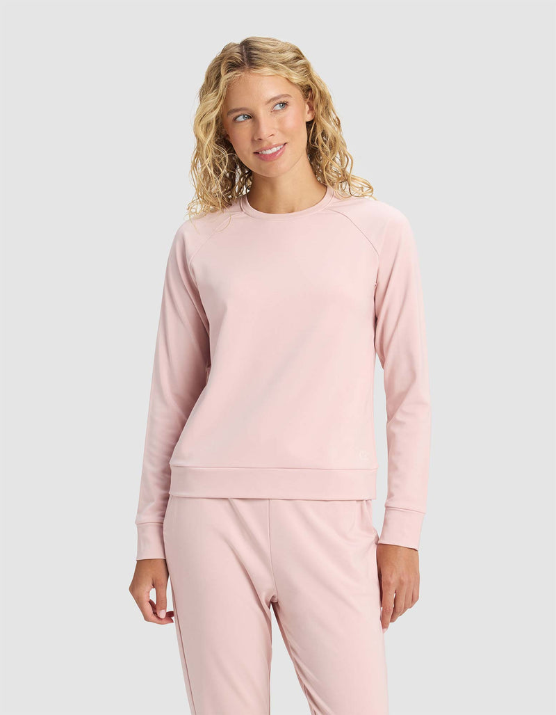 A woman with blonde, wavy hair wears the Cozy Earth Women's Brushed Bamboo Pullover Crew in light pink, paired with matching pants, standing against a plain light gray background and smiling softly while looking to the side.