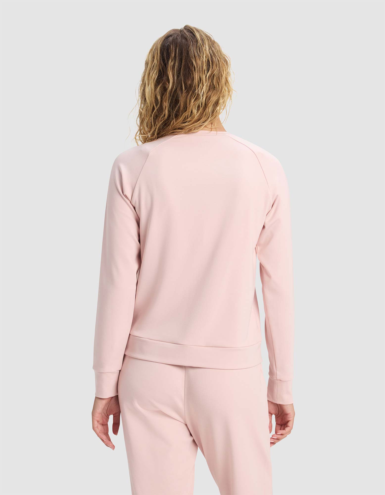 A woman with wavy, shoulder-length hair is seen from behind wearing the Cozy Earth Women's Brushed Bamboo Pullover Crew in light pink, paired with matching pants, standing against a plain gray background.