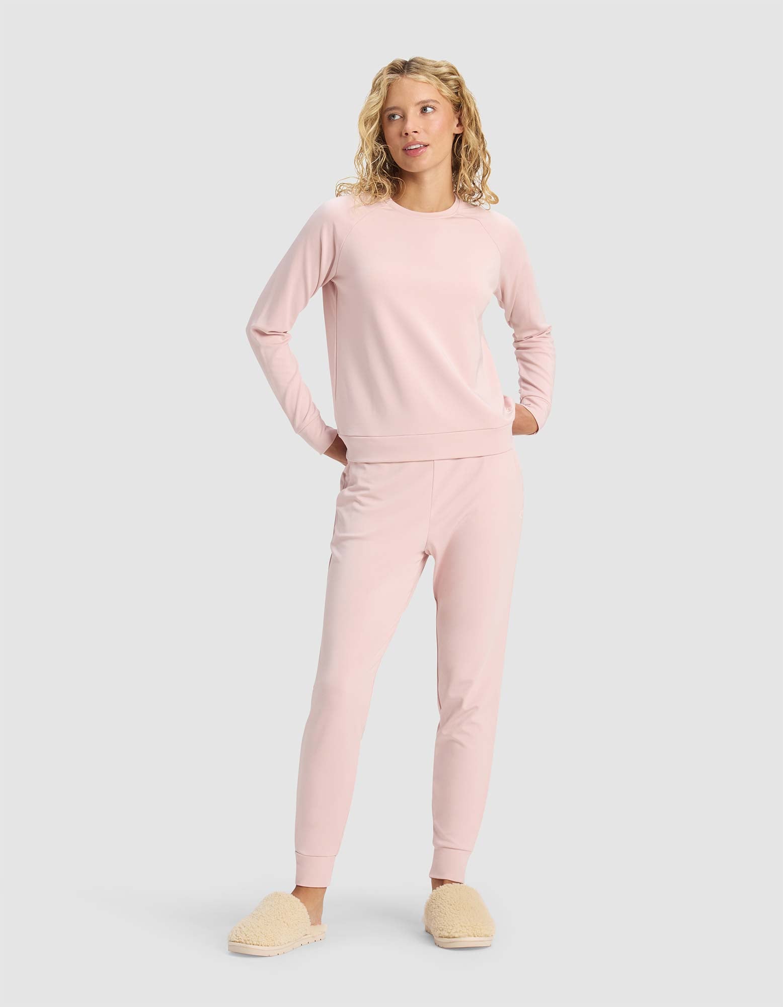 A person stands with hands in pockets, wearing the Cozy Earth Women's Brushed Bamboo Pullover Crew in light pink with matching joggers and beige fuzzy slippers, against a plain light gray background.