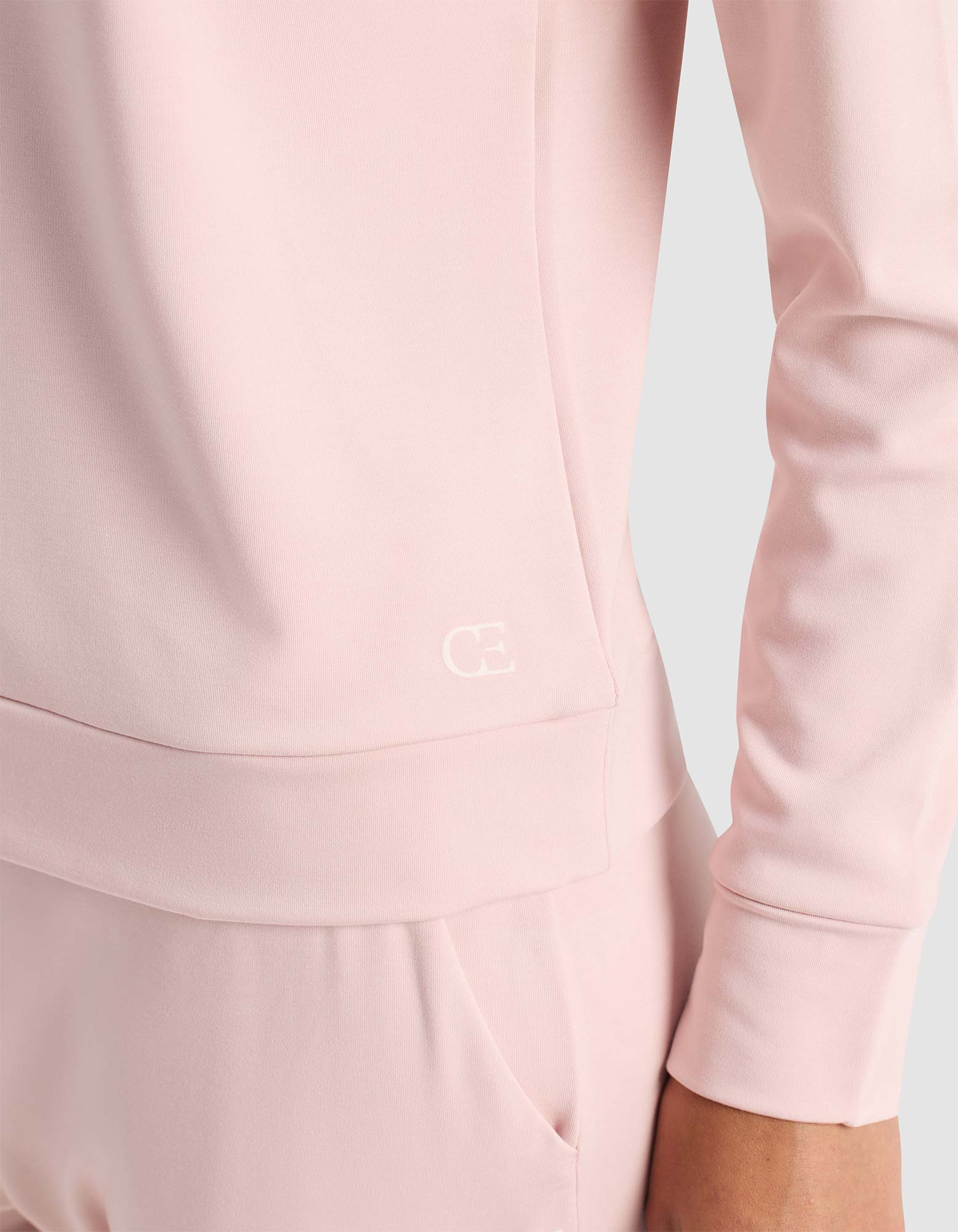 A woman wears the Cozy Earth Women's Brushed Bamboo Pullover Crew in light pink with matching pants, featuring a smooth fabric and a small white "CE" logo on the lower side of the top. One hand rests in her pocket.