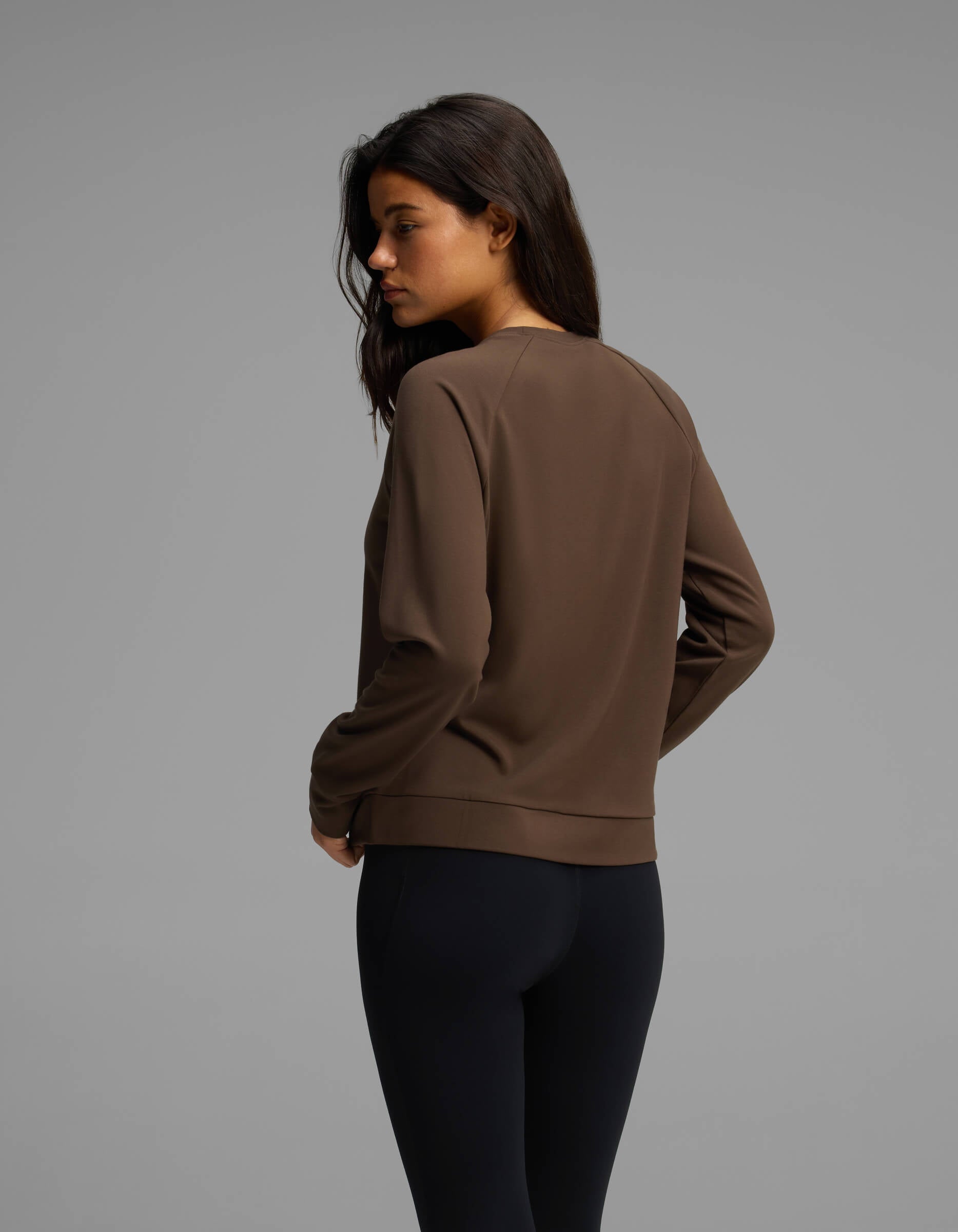 A woman with long dark hair, dressed in the Cozy Earth Women's Brushed Bamboo Pullover Crew and black leggings, stands turned slightly away from the camera against a plain gray background.|Color:Espresso