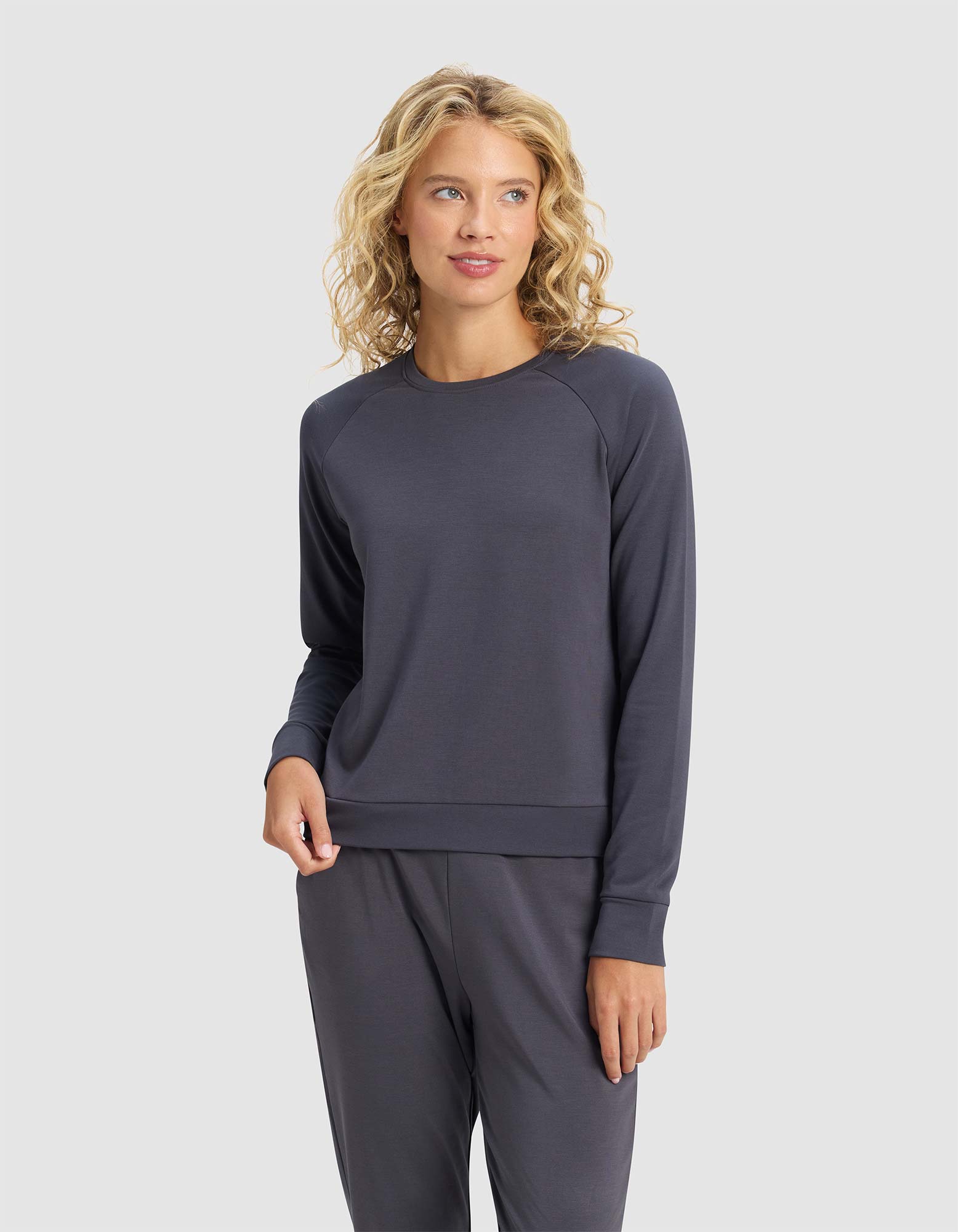 A woman with curly blonde hair wears the Cozy Earth Women's Brushed Bamboo Pullover Crew in dark gray, standing against a plain light gray background.