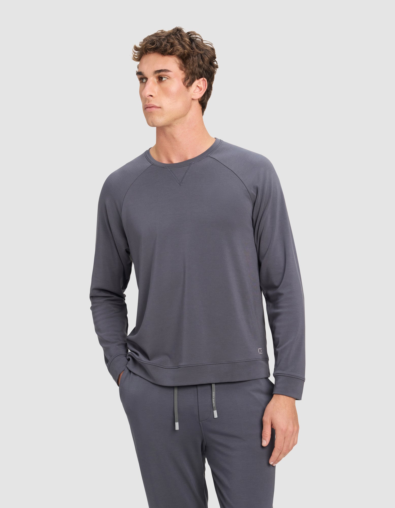 A man wears the Cozy Earth Men's Brushed Bamboo Pullover Crew in gray, paired with matching jogger pants, standing against a plain light background and looking slightly left with one hand in his pocket.