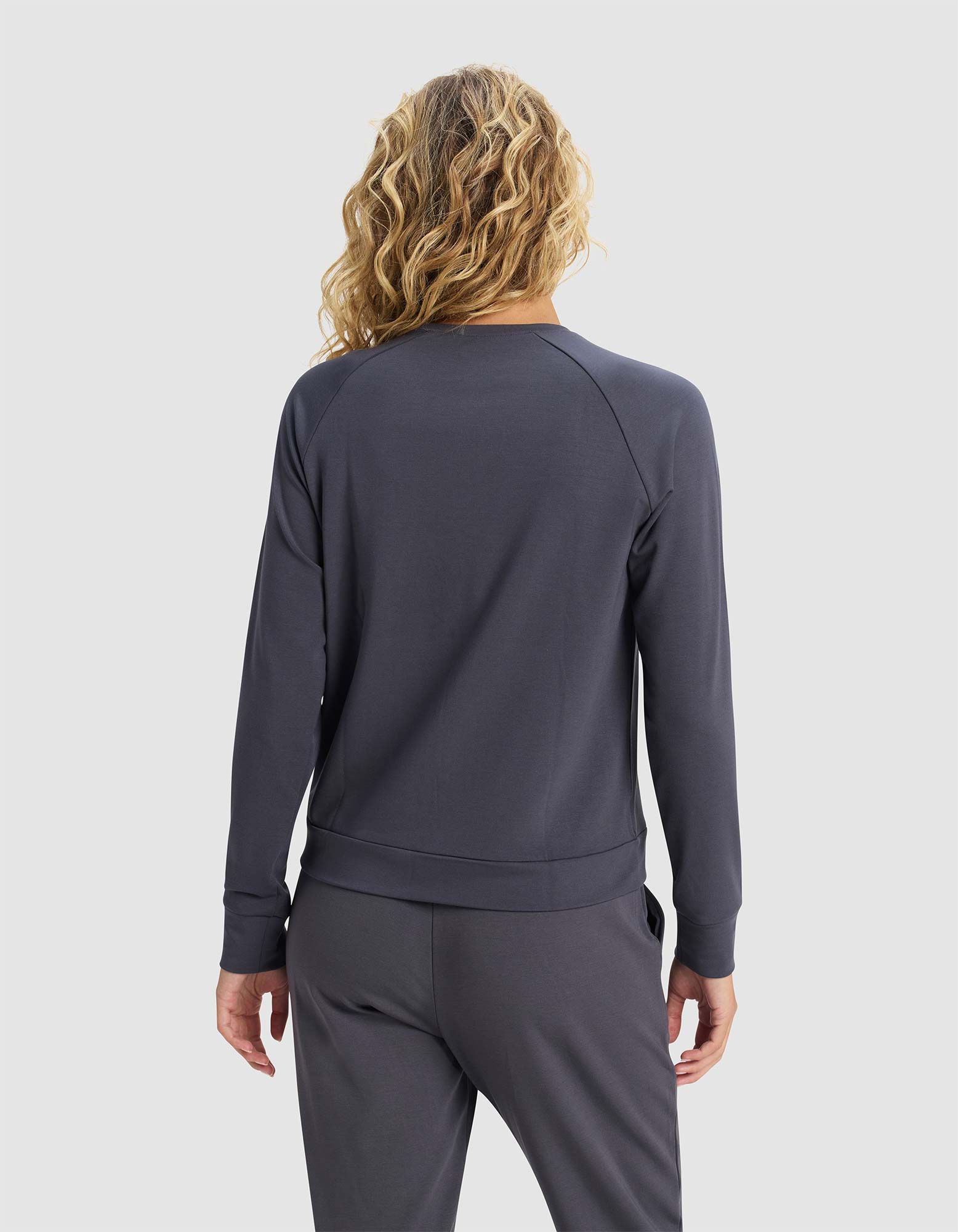 A person with wavy blonde hair stands facing away from the camera against a plain light background, wearing Cozy Earth's Women's Brushed Bamboo Pullover Crew in dark gray with matching pants.