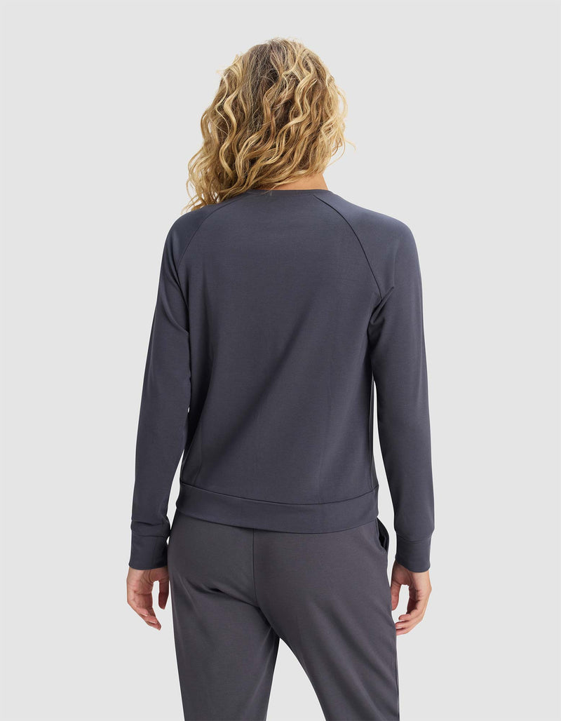 A person with wavy blonde hair stands facing away from the camera against a plain light background, wearing Cozy Earth's Women's Brushed Bamboo Pullover Crew in dark gray with matching pants.