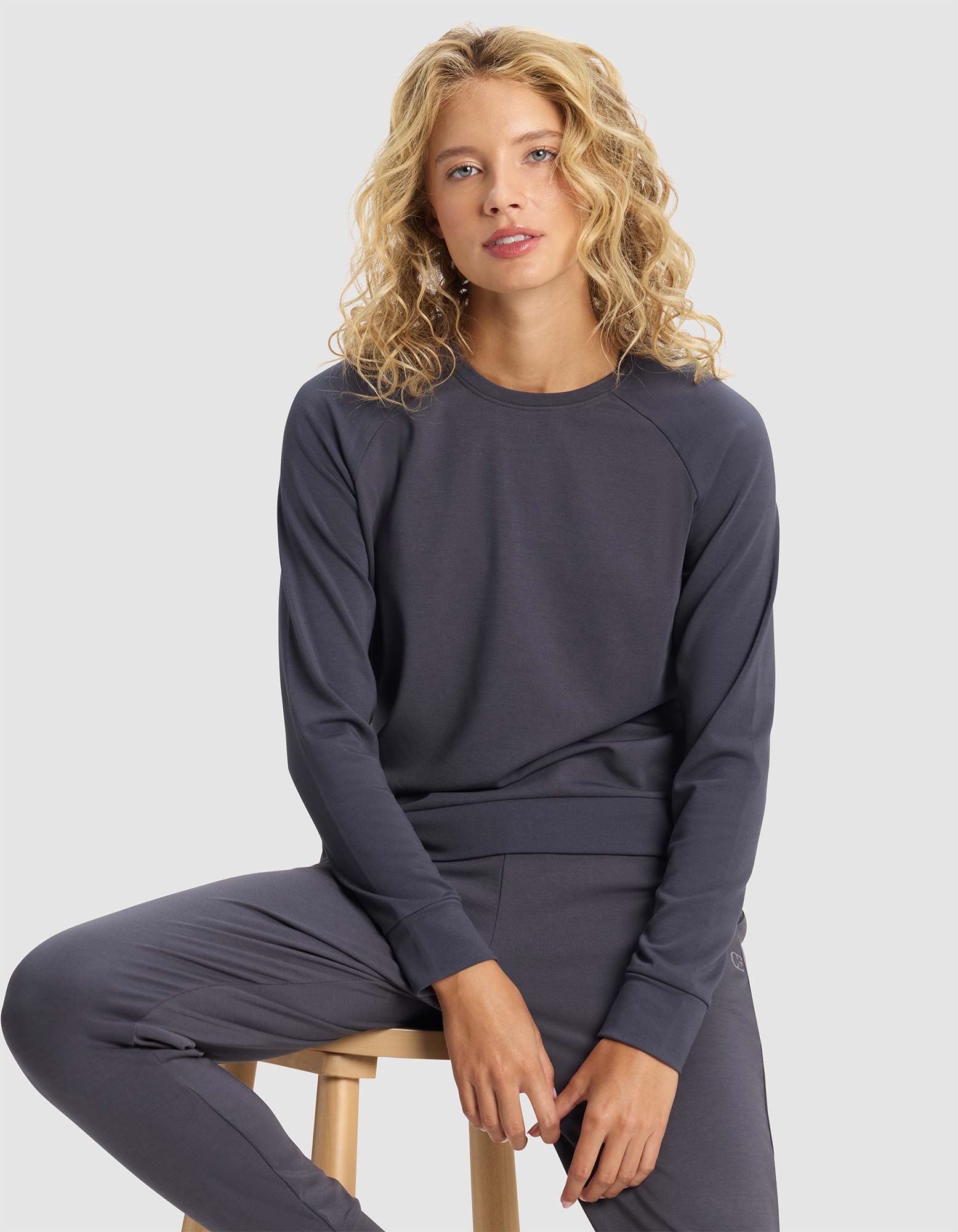 A woman with blonde curly hair wears the Cozy Earth Women's Brushed Bamboo Pullover Crew in dark gray while sitting on a wooden stool against a plain light gray background.