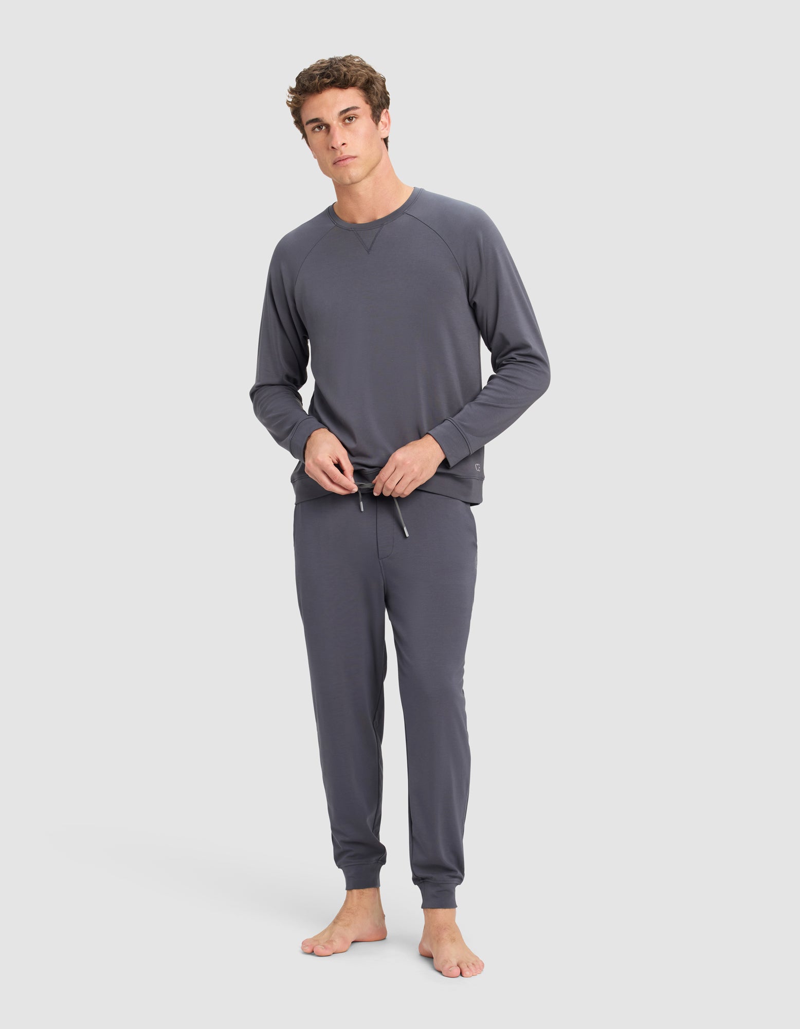 A young man stands barefoot against a plain background, wearing Cozy Earth's Men's Brushed Bamboo Pullover Crew with jogger-style pants. He looks at the camera while adjusting the drawstring on his pants.
