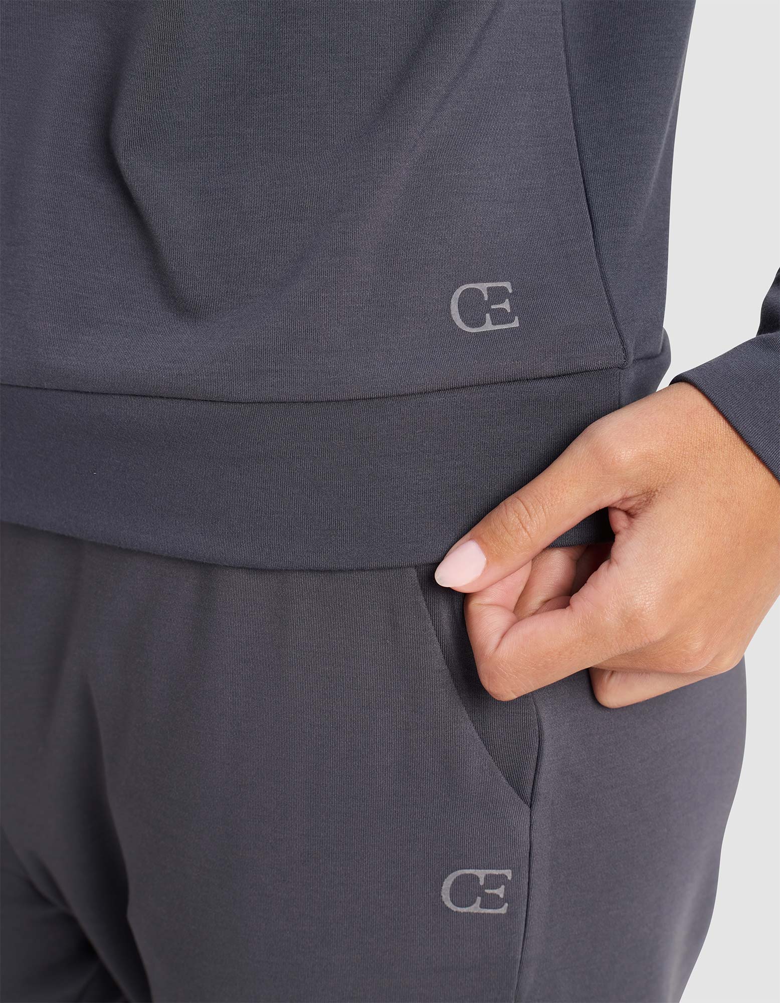 A person wears the Cozy Earth Women's Brushed Bamboo Pullover Crew in dark gray, with a subtle "CE" logo and their right hand in the pants pocket, paired with matching bottoms.