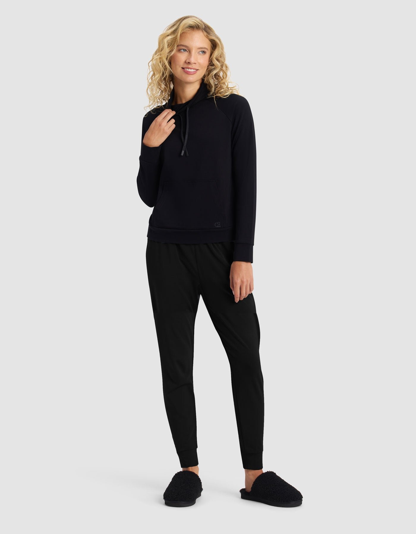 A woman with curly blonde hair smiles, standing against a light gray background, wearing the Cozy Earth Women's Brushed Bamboo Mock Neck & Jogger Set in black. She has one hand near her chin.