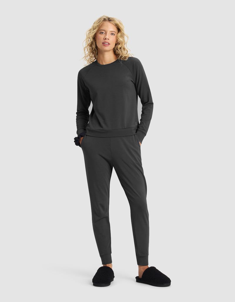 A woman with long, curly blond hair models the Cozy Earth Women's Brushed Bamboo Jogger Set in dark gray and black slippers, standing hands in pockets against a plain light gray background.