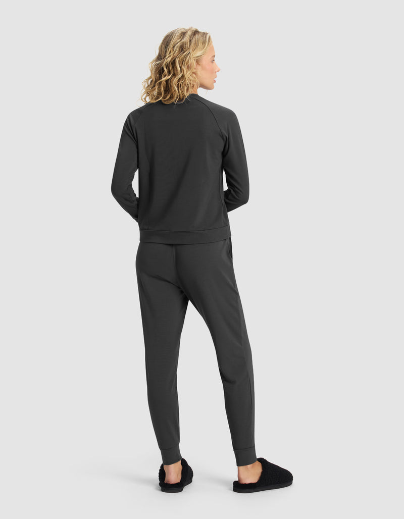 A woman with blonde, wavy hair faces away, wearing the Cozy Earth Women's Brushed Bamboo Jogger Set in dark gray with black slippers, against a plain light gray background.