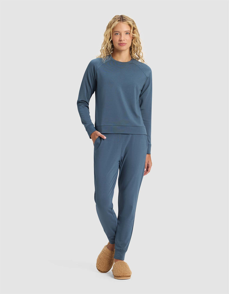 A woman with long curly blonde hair models the Cozy Earth Women's Brushed Bamboo Jogger Set in blue, standing with one hand in her pocket and wearing brown fuzzy slippers against a plain light background.
