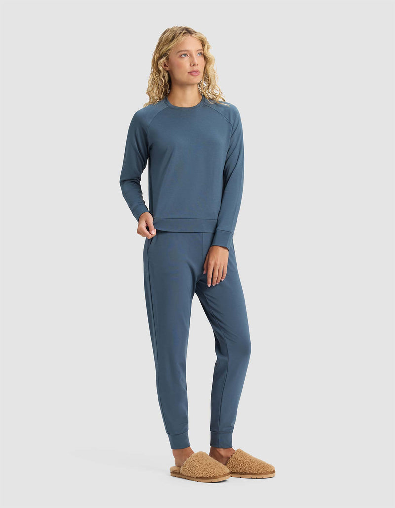 A woman with curly blonde hair wears the Cozy Earth Women's Brushed Bamboo Jogger Set in blue with tan fuzzy slippers, standing against a plain light background.