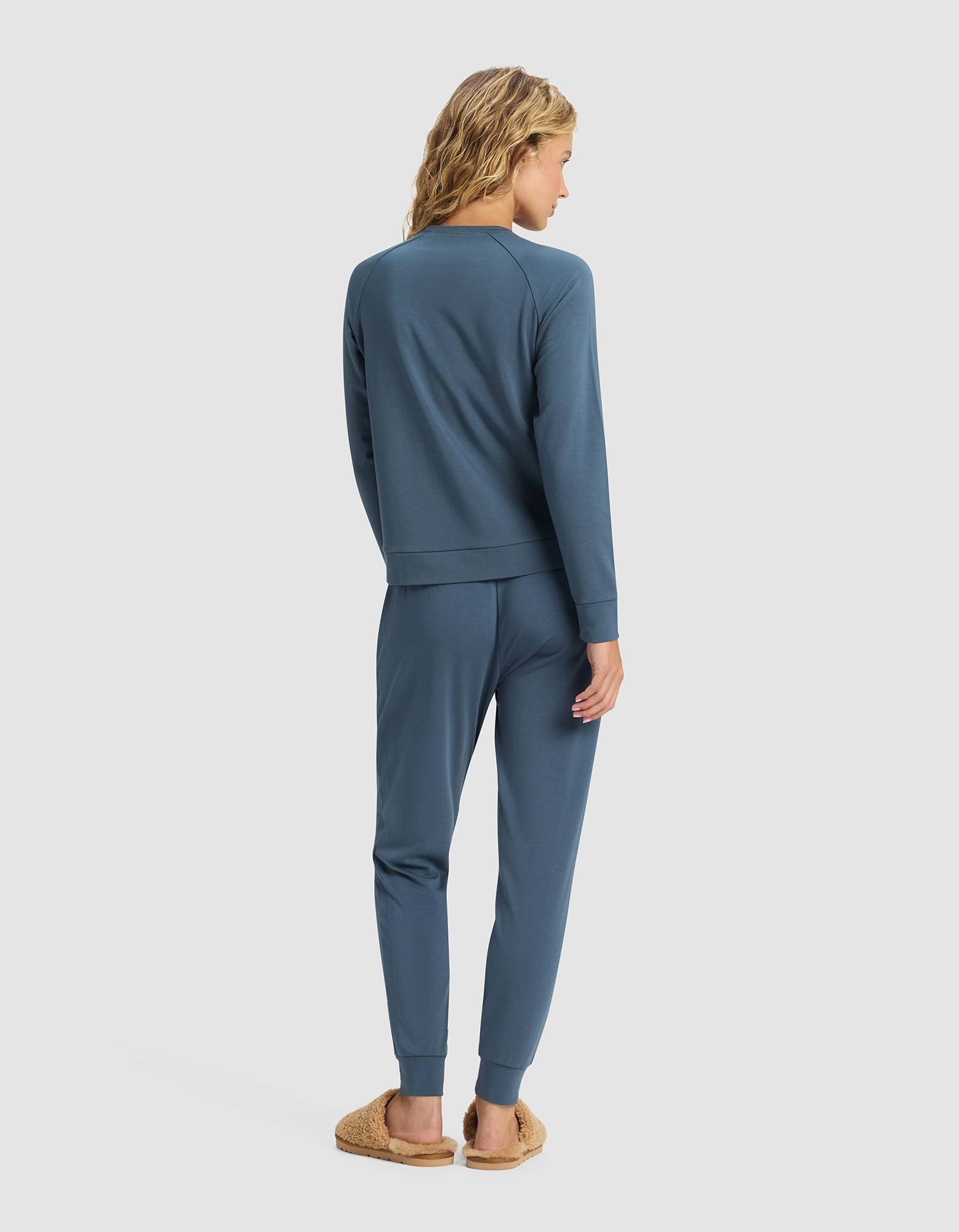 A person with wavy blonde hair stands facing away, wearing the Cozy Earth Women's Brushed Bamboo Jogger Set in blue and tan fuzzy slippers, set against a plain light background.