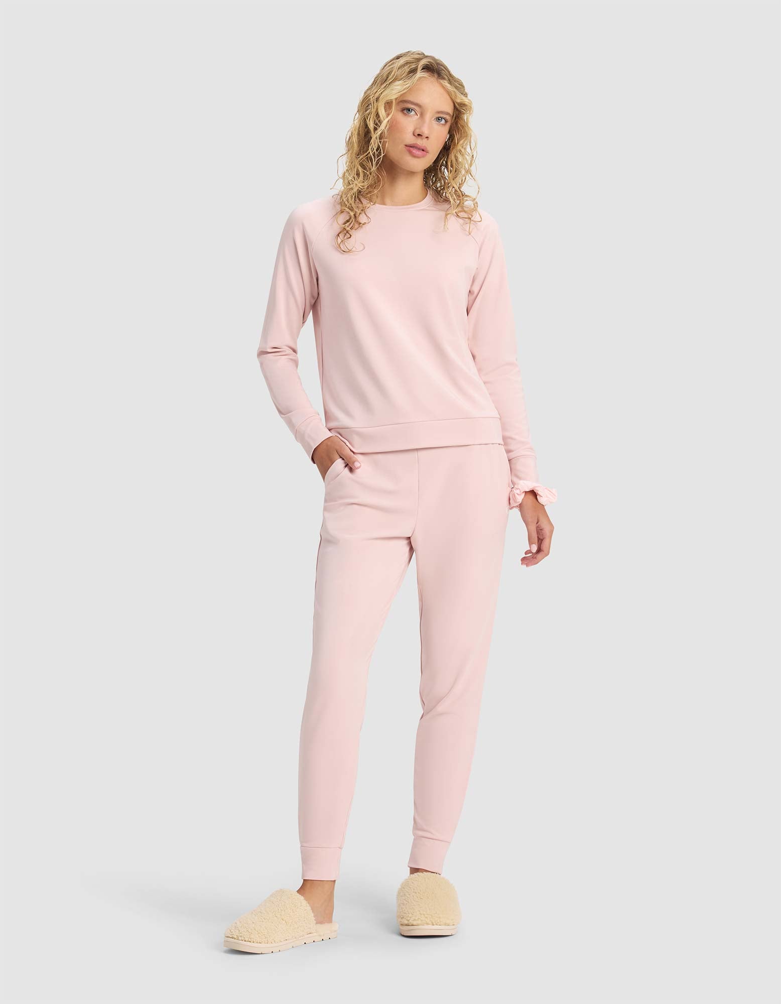 A woman with long, curly blonde hair wears the Cozy Earth Women's Brushed Bamboo Jogger Set in light pink with beige fuzzy slippers, standing against a plain light gray background.