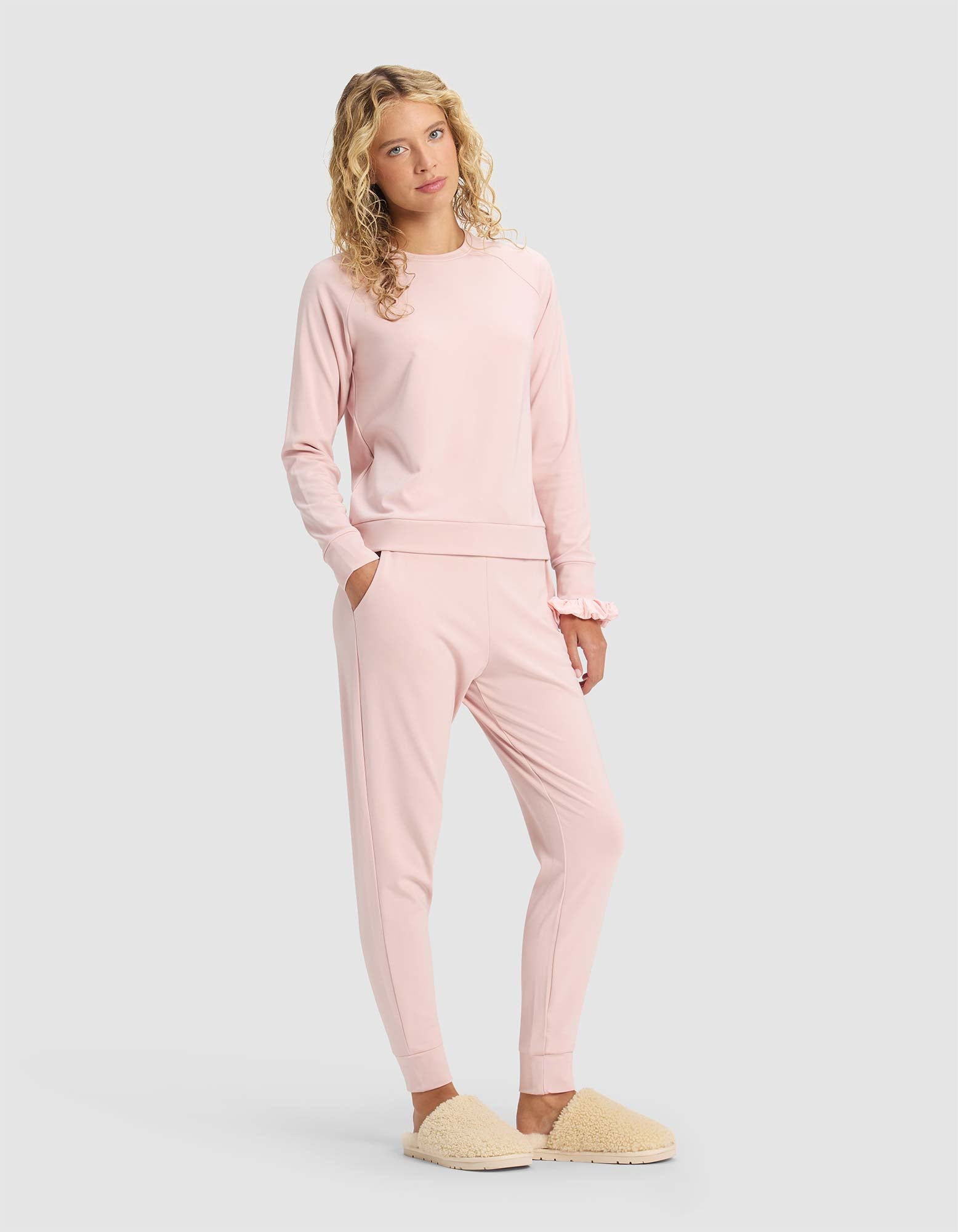 A young woman with curly blonde hair models the Cozy Earth Women's Brushed Bamboo Jogger Set in light pink, paired with beige fuzzy slippers, standing against a plain light gray background with one hand in her pocket.