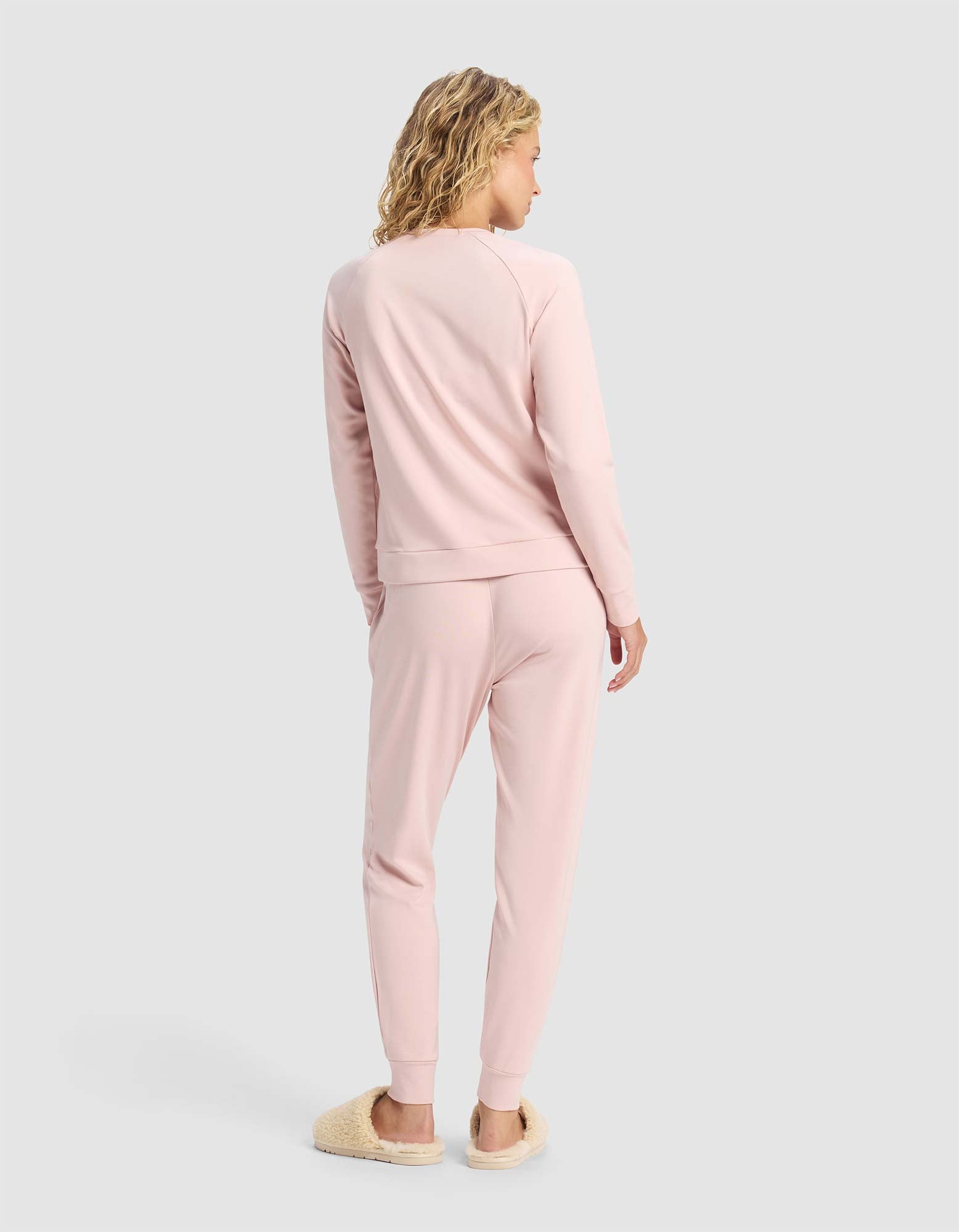 A woman with curly blonde hair faces away, wearing the Cozy Earth Women's Brushed Bamboo Jogger Set in light pink and beige fuzzy slippers, set against a plain light gray background.