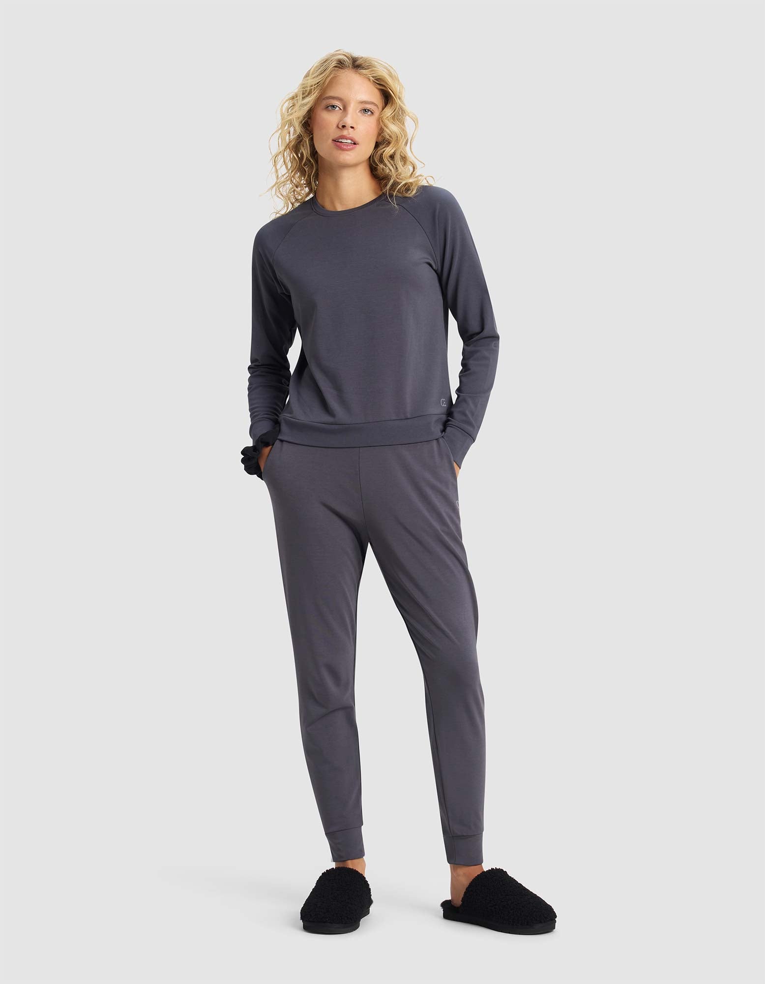 A woman with blonde curly hair wears the Cozy Earth Women's Brushed Bamboo Jogger Set in dark grey, with her hands in her pockets and black fuzzy slippers, standing against a plain light background.