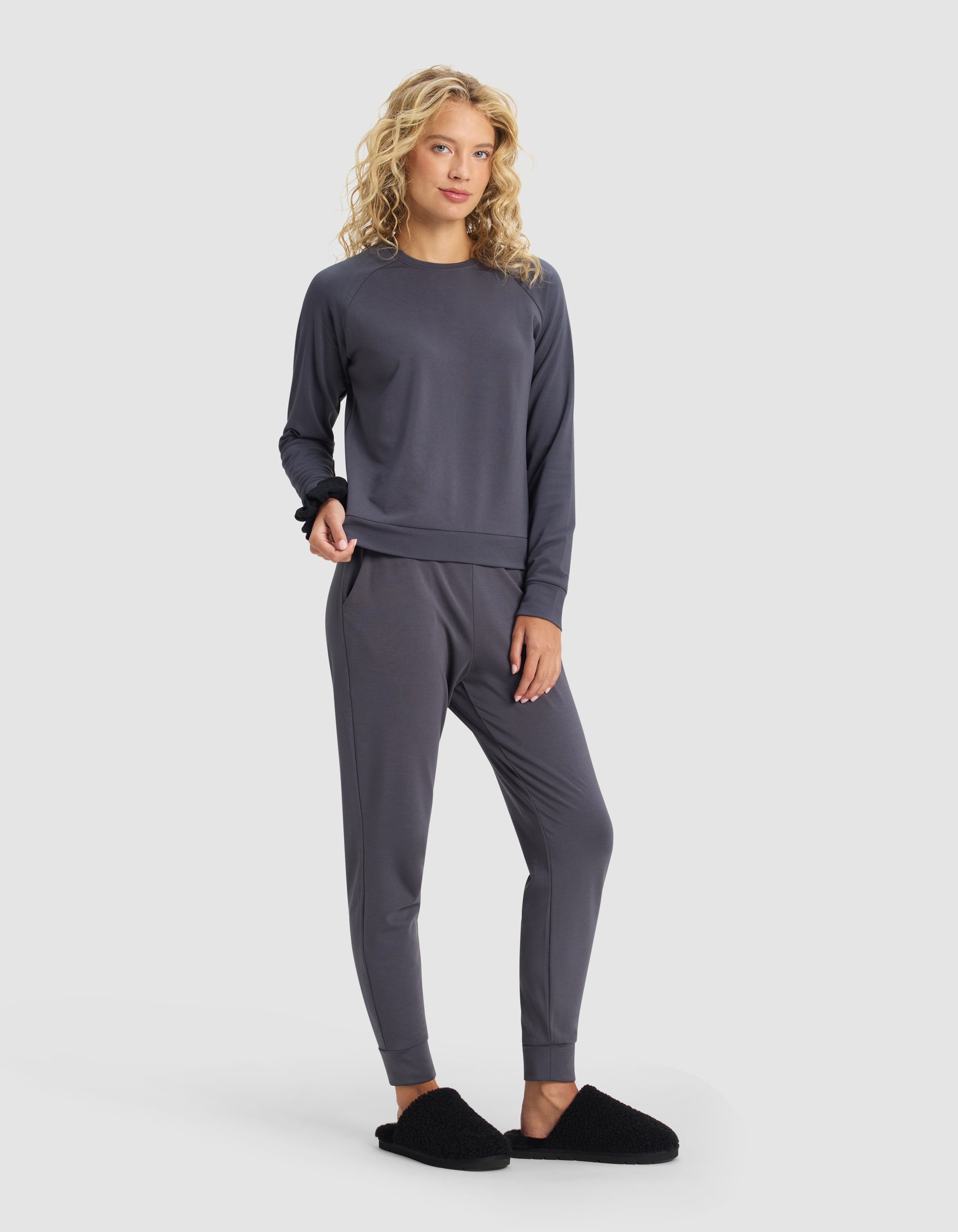 A woman with long curly blonde hair models the Cozy Earth Women's Brushed Bamboo Jogger Set in charcoal gray, paired with black slippers, standing against a plain light gray background.