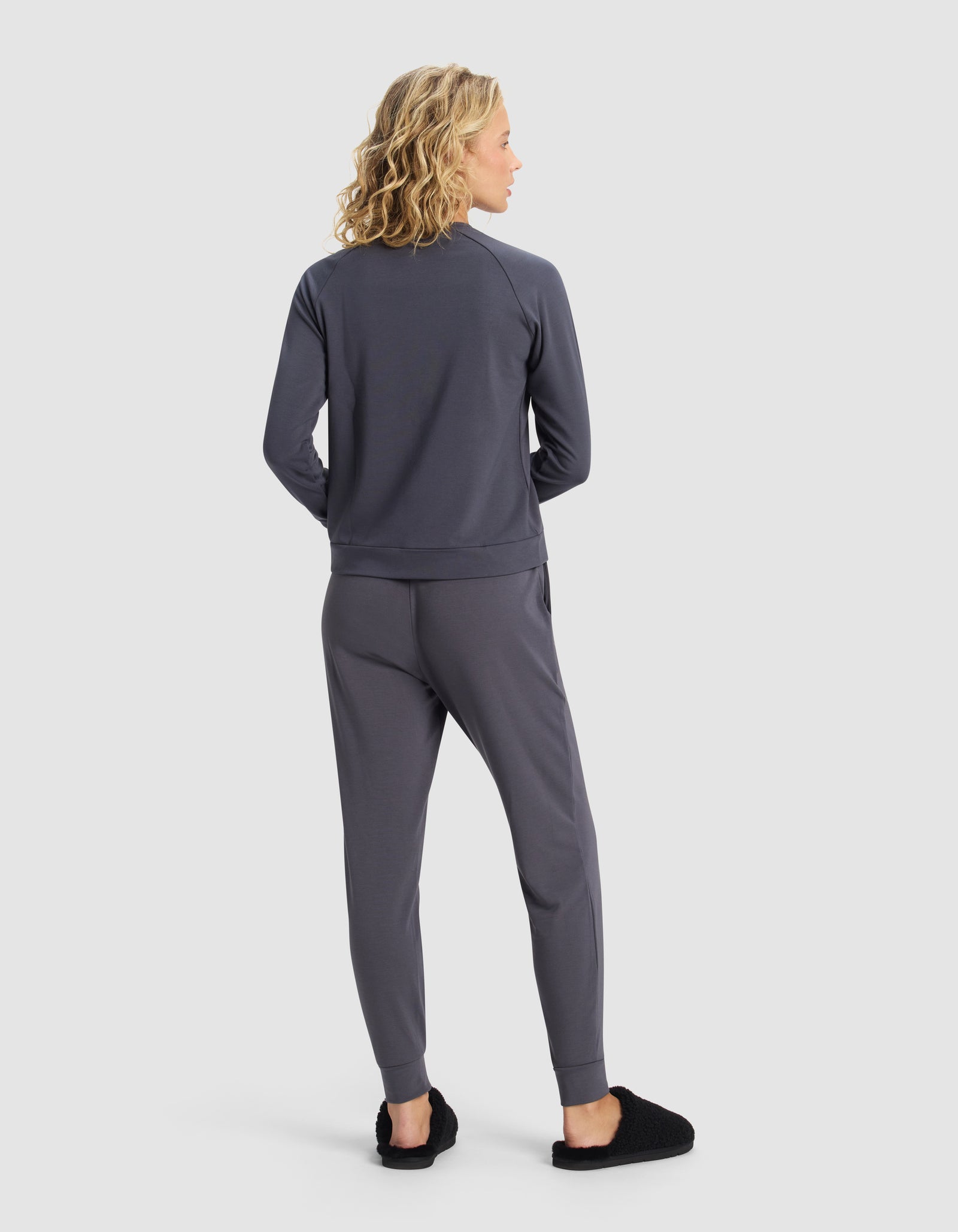 A woman with wavy blonde hair faces away, wearing the Cozy Earth Women's Brushed Bamboo Jogger Set in dark gray and black slippers, set against a plain white background.