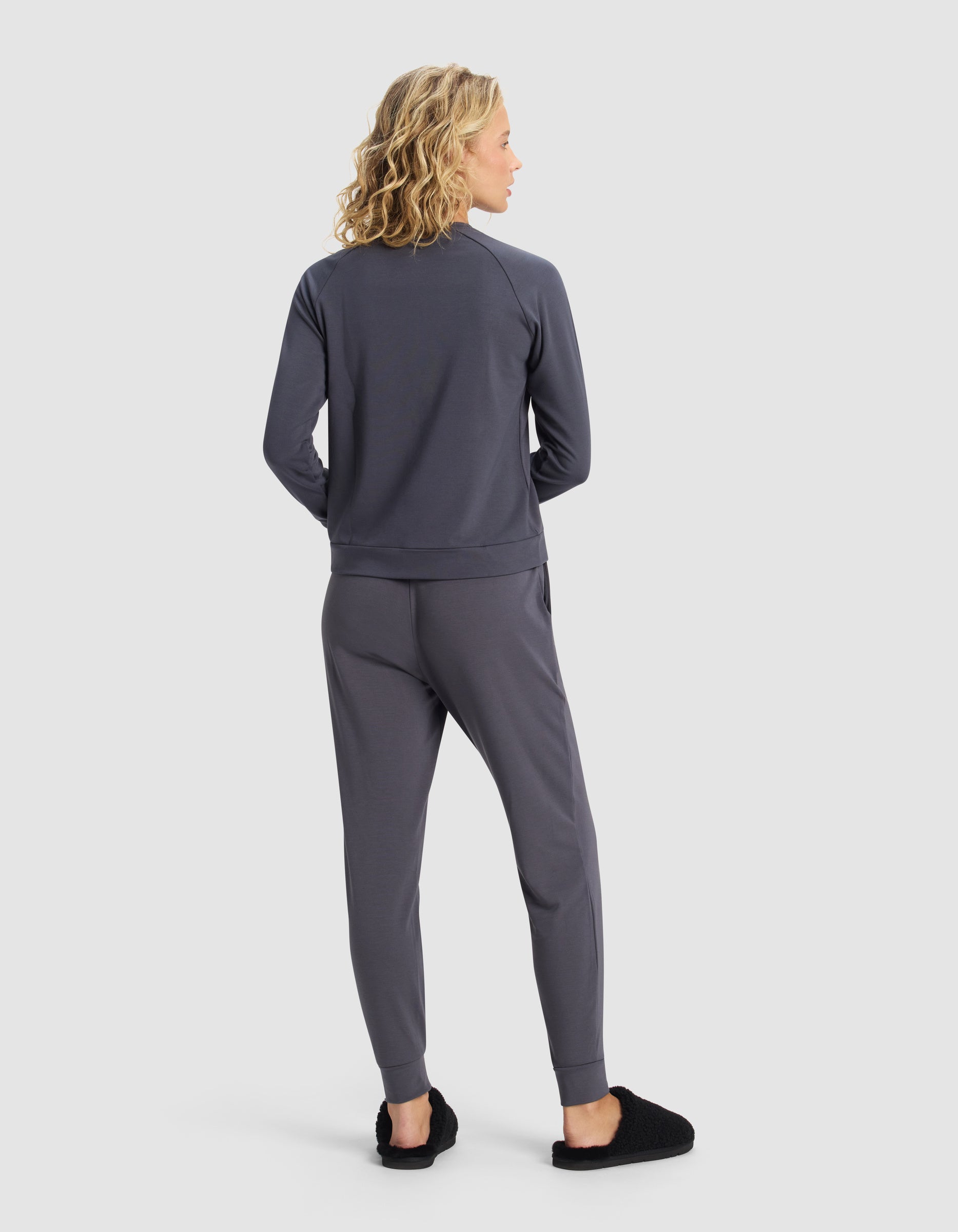 A woman with wavy blonde hair faces away, wearing the Cozy Earth Women's Brushed Bamboo Jogger Set in dark gray and black slippers, set against a plain white background.