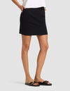 A person stands with hands in pockets against a plain light background, wearing Cozy Earth's Women's Brushed Bamboo Skirt in black, a white top, and black slide sandals with gold buckles. |Color:Black