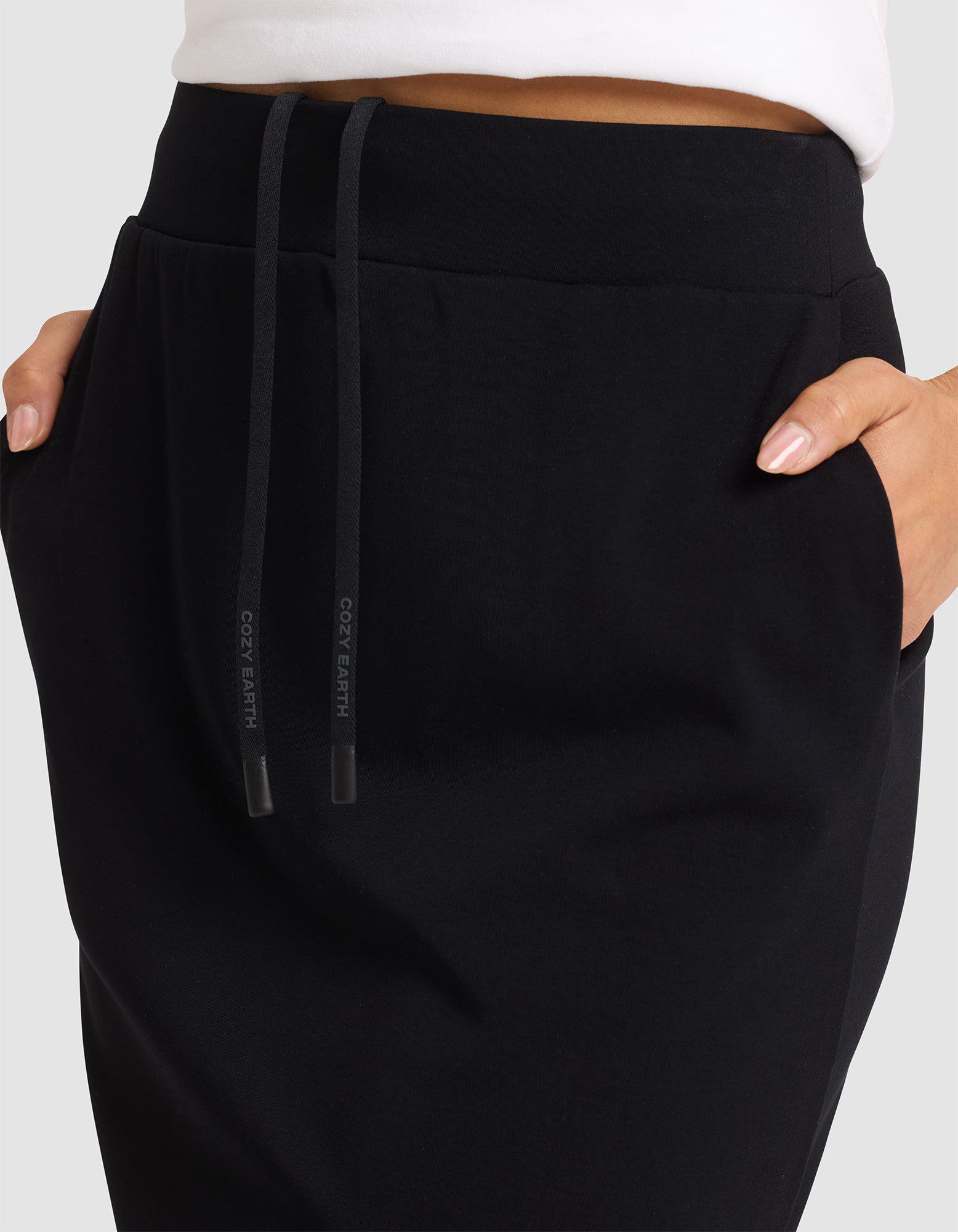 A person is shown from the waist down wearing the Cozy Earth Women's Brushed Bamboo Skirt in black, with hands in pockets, against a plain background. 