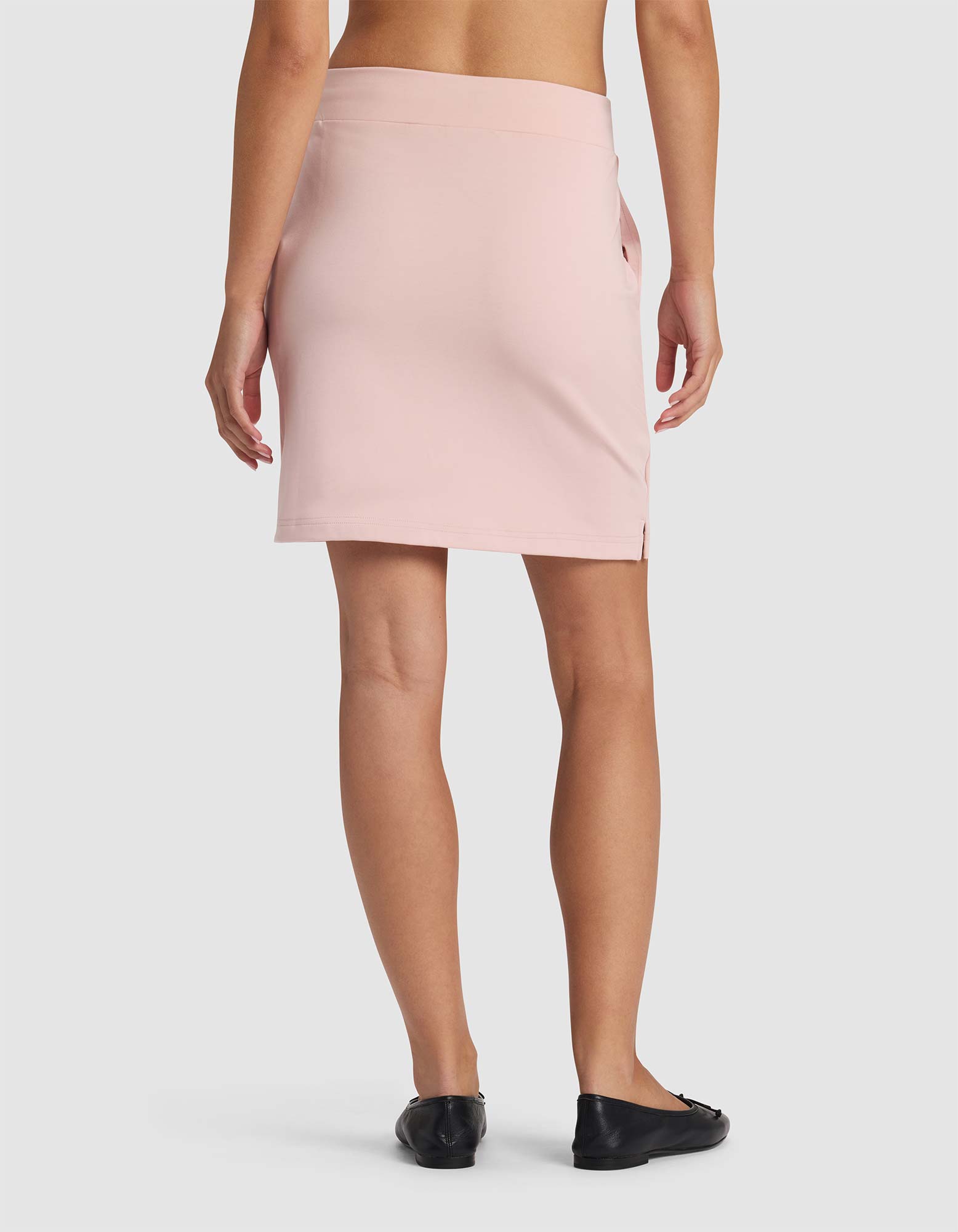 A person is seen from behind wearing Cozy Earth's Women's Brushed Bamboo Skirt in light pink with black flats, set against a plain white background.