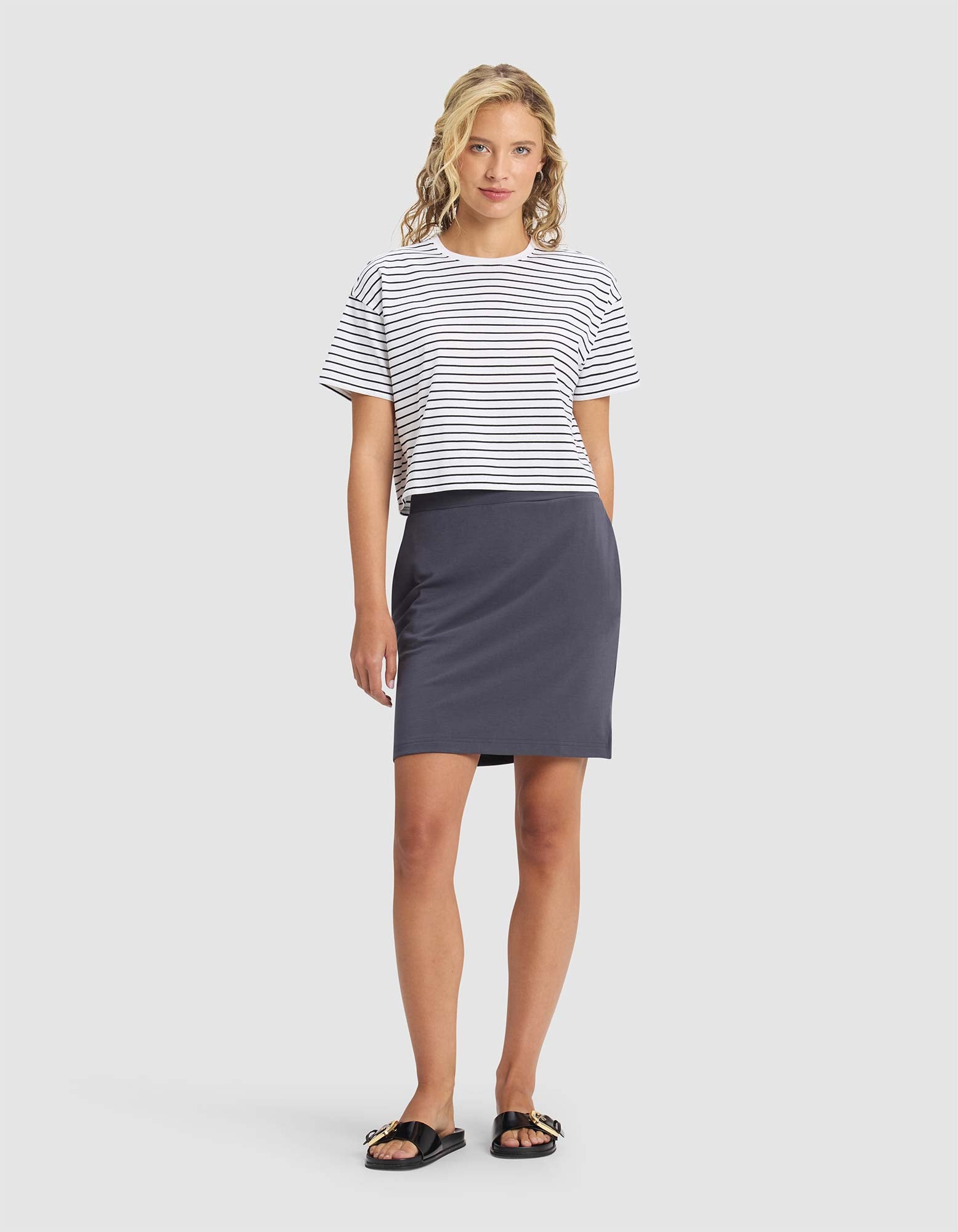 A woman with wavy blonde hair and a relaxed expression stands against a plain background wearing the Cozy Earth Women's Brushed Bamboo Skirt, paired with a black and white striped t-shirt and black sandals with gold chain details. 