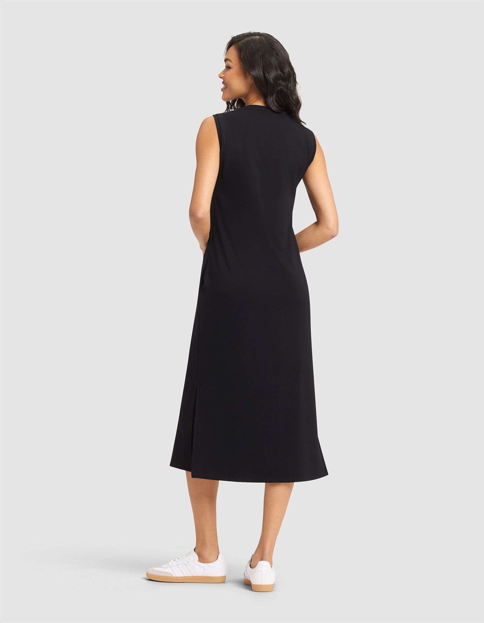 A woman with dark, wavy hair smiles while wearing the Cozy Earth Women's Brushed Bamboo Sleeveless Midi Dress in black and white sneakers, standing against a plain light gray background. |Color:Black