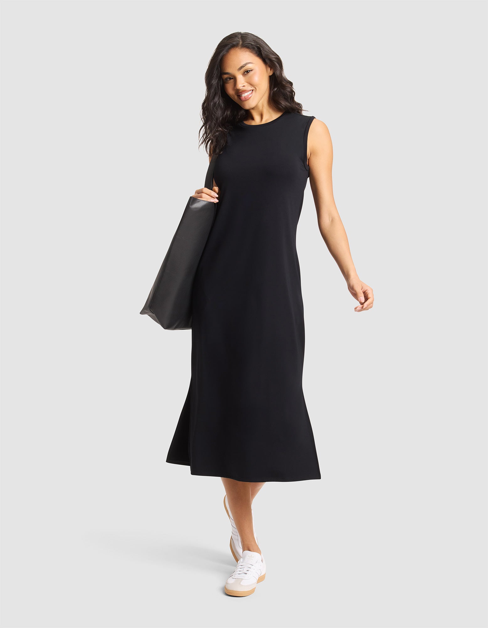 A woman smiles against a light gray background wearing Cozy Earth's Women's Brushed Bamboo Sleeveless Midi Dress in black, paired with white sneakers and a large black tote bag. 