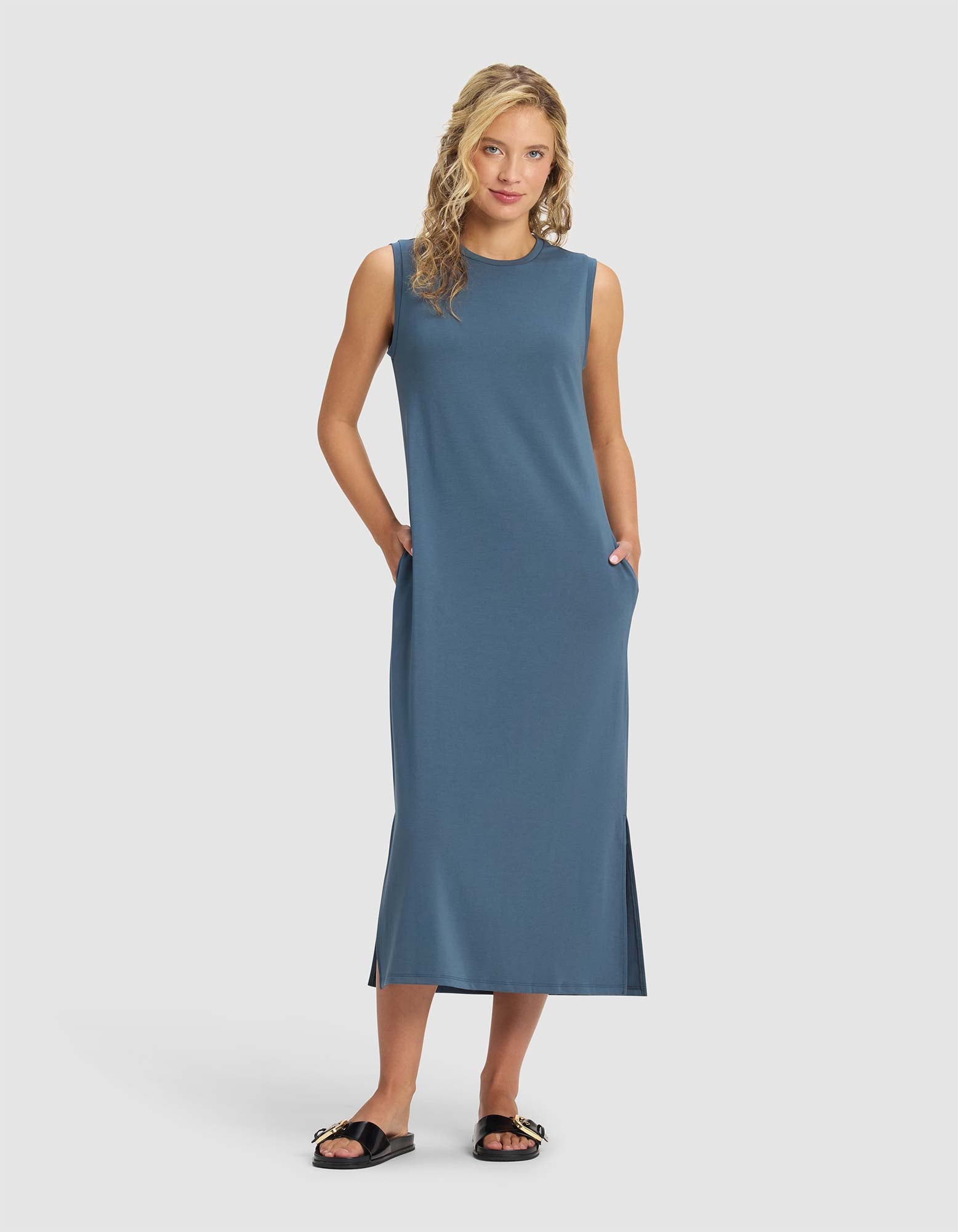 A woman with wavy blonde hair wears the Cozy Earth Women's Brushed Bamboo Sleeveless Midi Dress and black sandals, standing with her hands in her pockets against a plain light background. |Color:Dawn