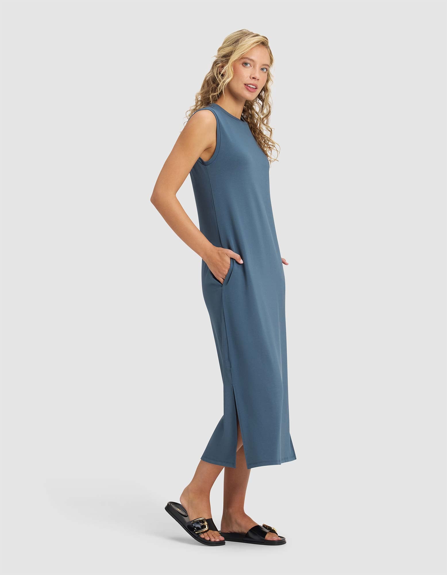 A woman with blonde hair wears the Cozy Earth Women's Brushed Bamboo Sleeveless Midi Dress in blue with side pockets and black slide sandals, standing sideways and smiling against a plain white background.|Color:Dawn