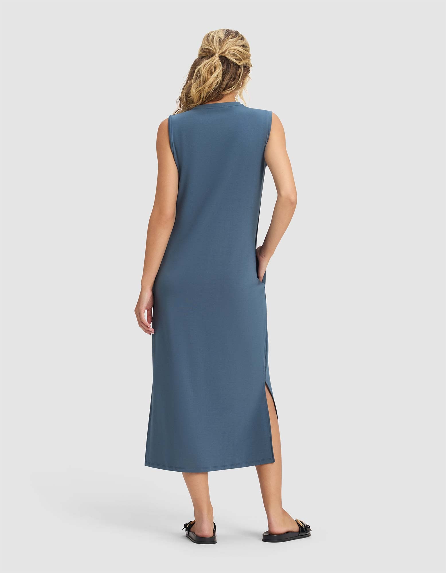 A woman faces away wearing the Cozy Earth Women's Brushed Bamboo Sleeveless Midi Dress in blue with a side slit, her hair half tied back, and black slide sandals. She stands against a plain, light background.|Color:Dawn