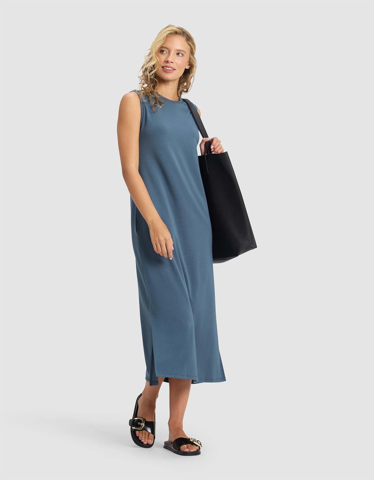 A woman stands against a plain light background in a Cozy Earth Women's Brushed Bamboo Sleeveless Midi Dress, paired with black slide sandals featuring gold accents and a large black tote bag.|Color:Dawn