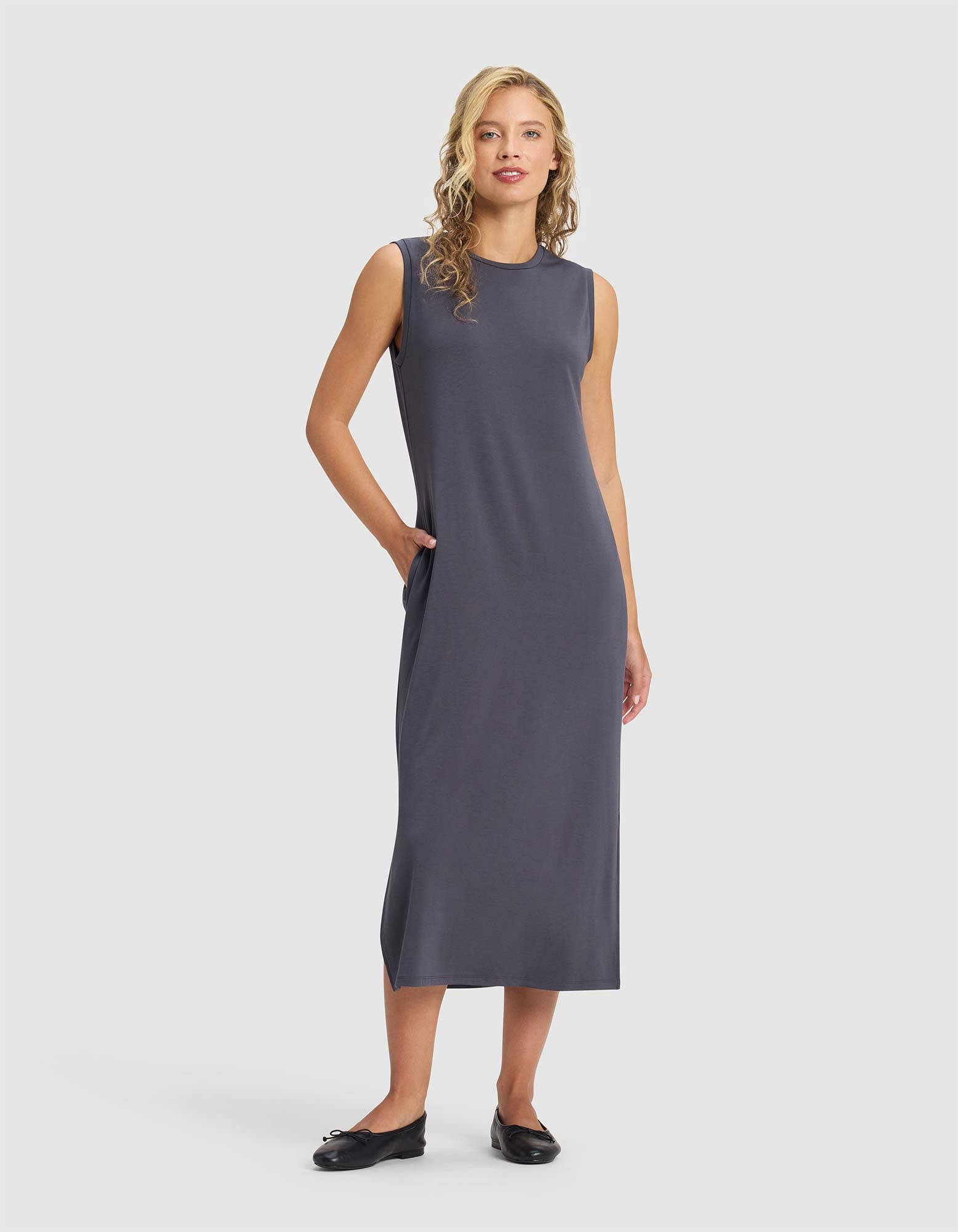 A woman with blonde curly hair wears the Cozy Earth Women's Brushed Bamboo Sleeveless Midi Dress in dark gray with black flats, standing against a plain light background, one hand in her pocket.