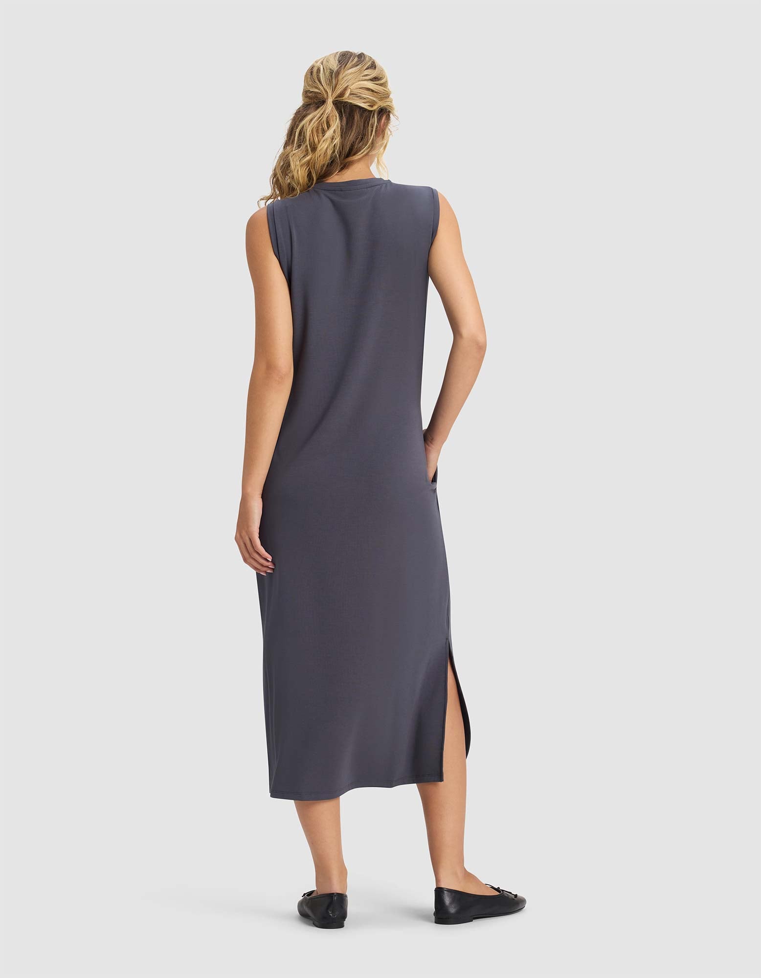 A woman with blonde hair in a ponytail faces away, wearing Cozy Earth's Women's Brushed Bamboo Sleeveless Midi Dress in dark gray with a side slit and black flats, against a plain white background.
