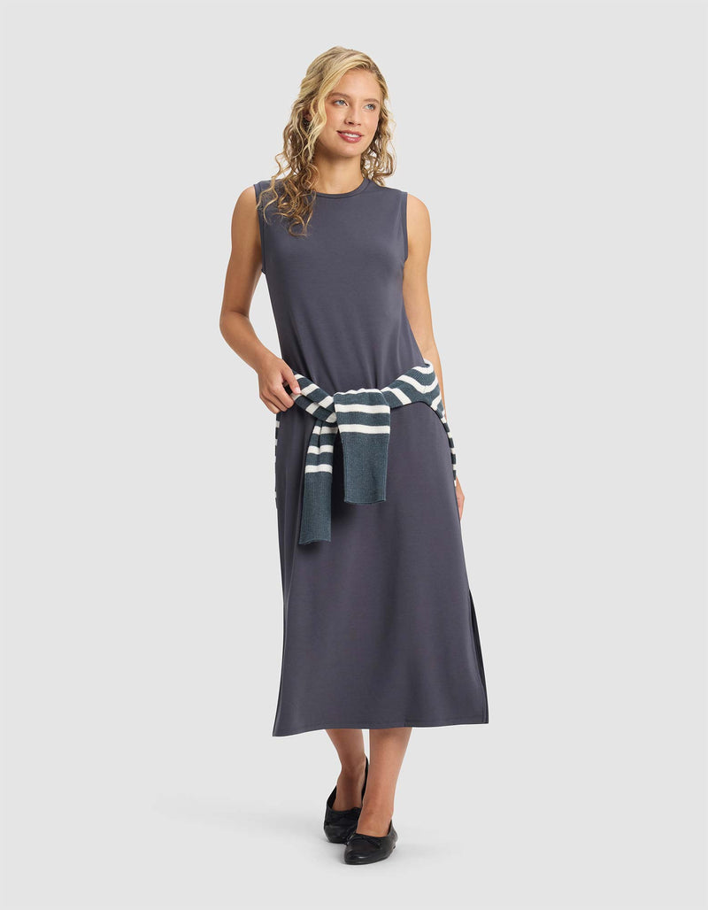 A woman with curly blonde hair smiles in front of a plain light background, wearing the Cozy Earth Women's Brushed Bamboo Sleeveless Midi Dress in dark gray with black shoes and a striped sweater tied at her waist.