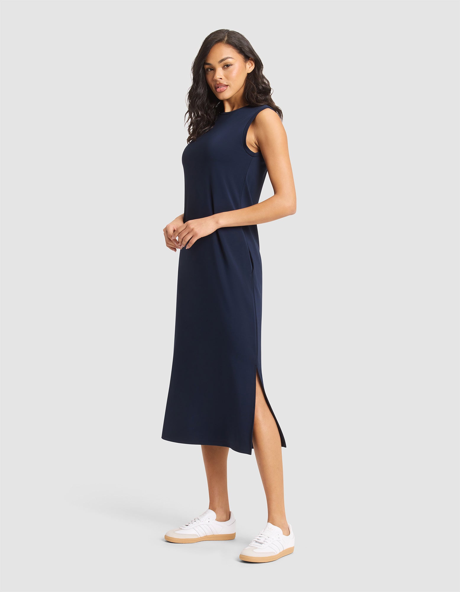 A woman in a Cozy Earth Women's Brushed Bamboo Sleeveless Midi Dress and white sneakers stands against a plain light gray background. 