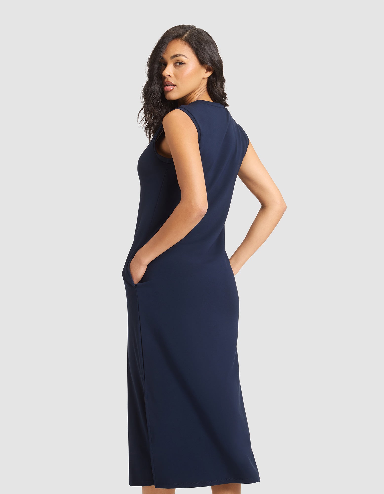 A woman with wavy dark hair models the Cozy Earth Women's Brushed Bamboo Sleeveless Midi Dress with pockets, in navy blue, standing sideways and looking over her shoulder against a plain light gray background. 