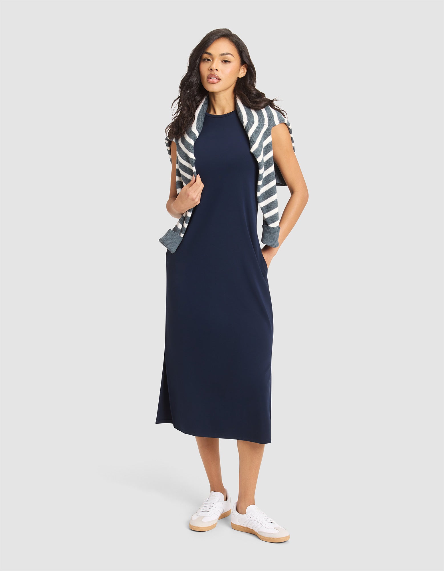A woman models the Cozy Earth Women's Brushed Bamboo Sleeveless Midi Dress in navy, styled with a striped sweater over her shoulders and white sneakers, posing with one hand in her pocket against a plain light background. 