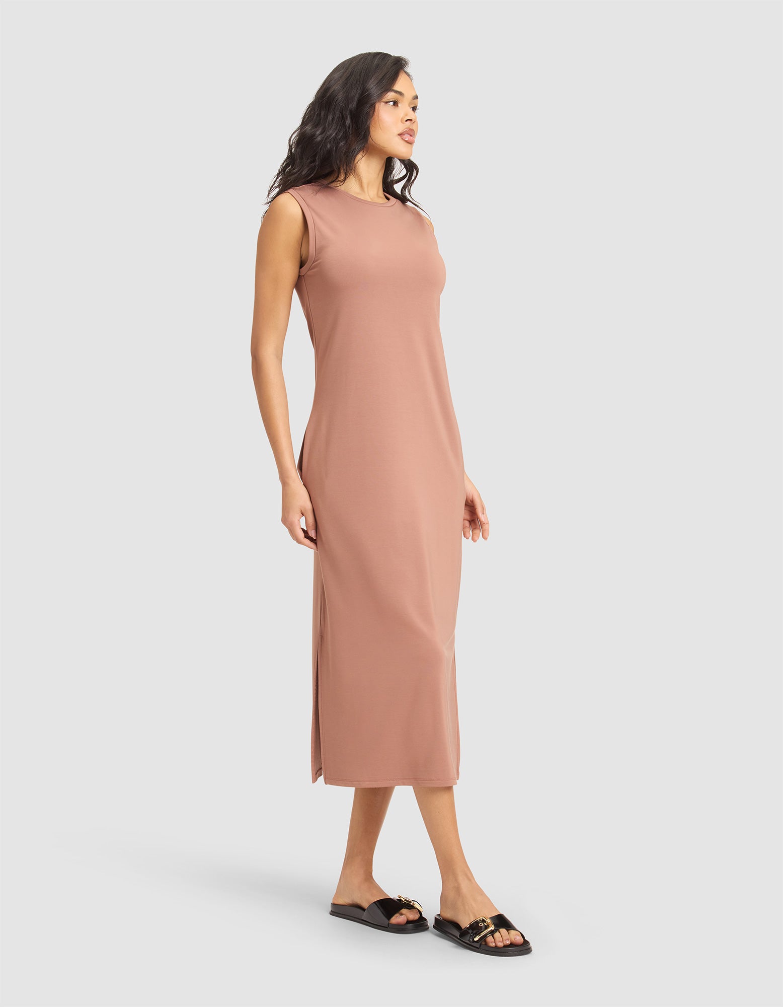 A woman with medium-length dark hair wears the Cozy Earth Women's Brushed Bamboo Sleeveless Midi Dress in light brown, featuring side slits and a midi length. She pairs it with black slide sandals against a plain white background. 