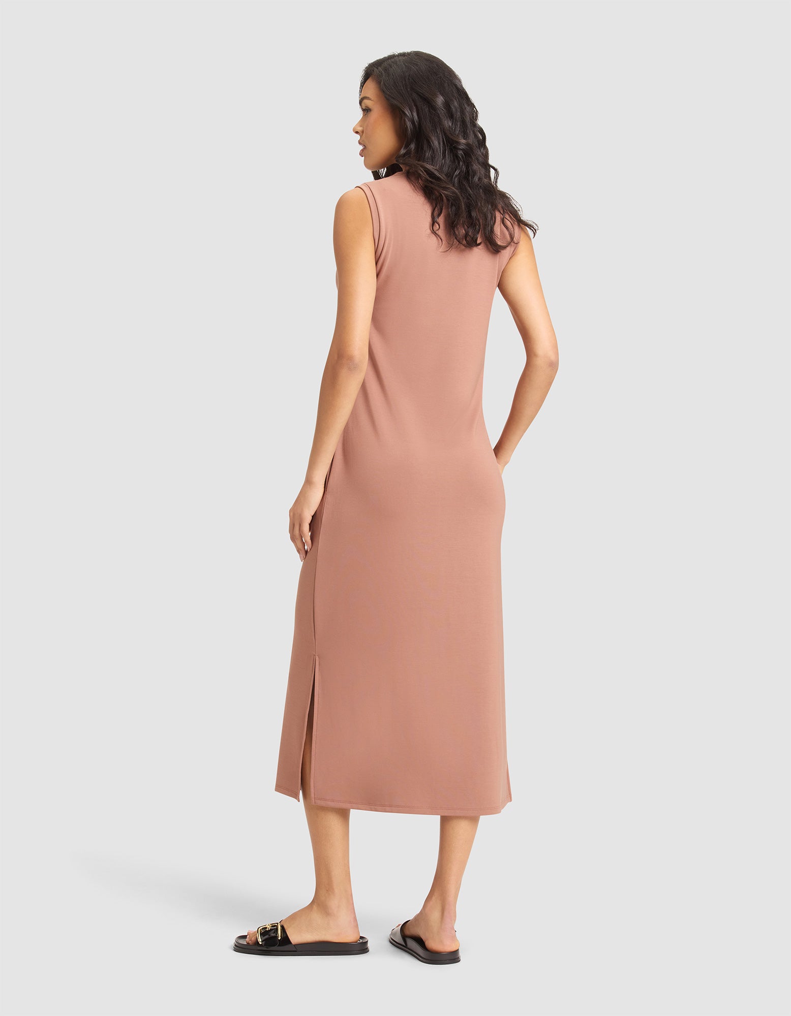 A woman with wavy dark hair, wearing black flat sandals and the Cozy Earth Women's Brushed Bamboo Sleeveless Midi Dress in rose, stands facing away against a plain light gray background. The knee-length dress features a side slit. 