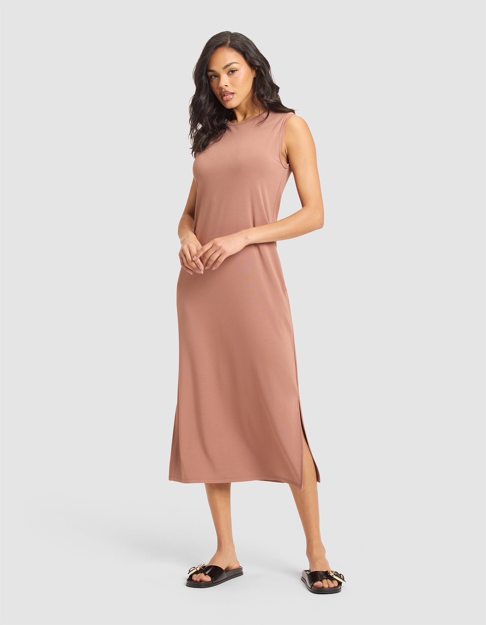 A woman models the Cozy Earth Women's Brushed Bamboo Sleeveless Midi Dress in light brown, paired with black slide sandals featuring gold accents, posing against a plain light gray background. 
