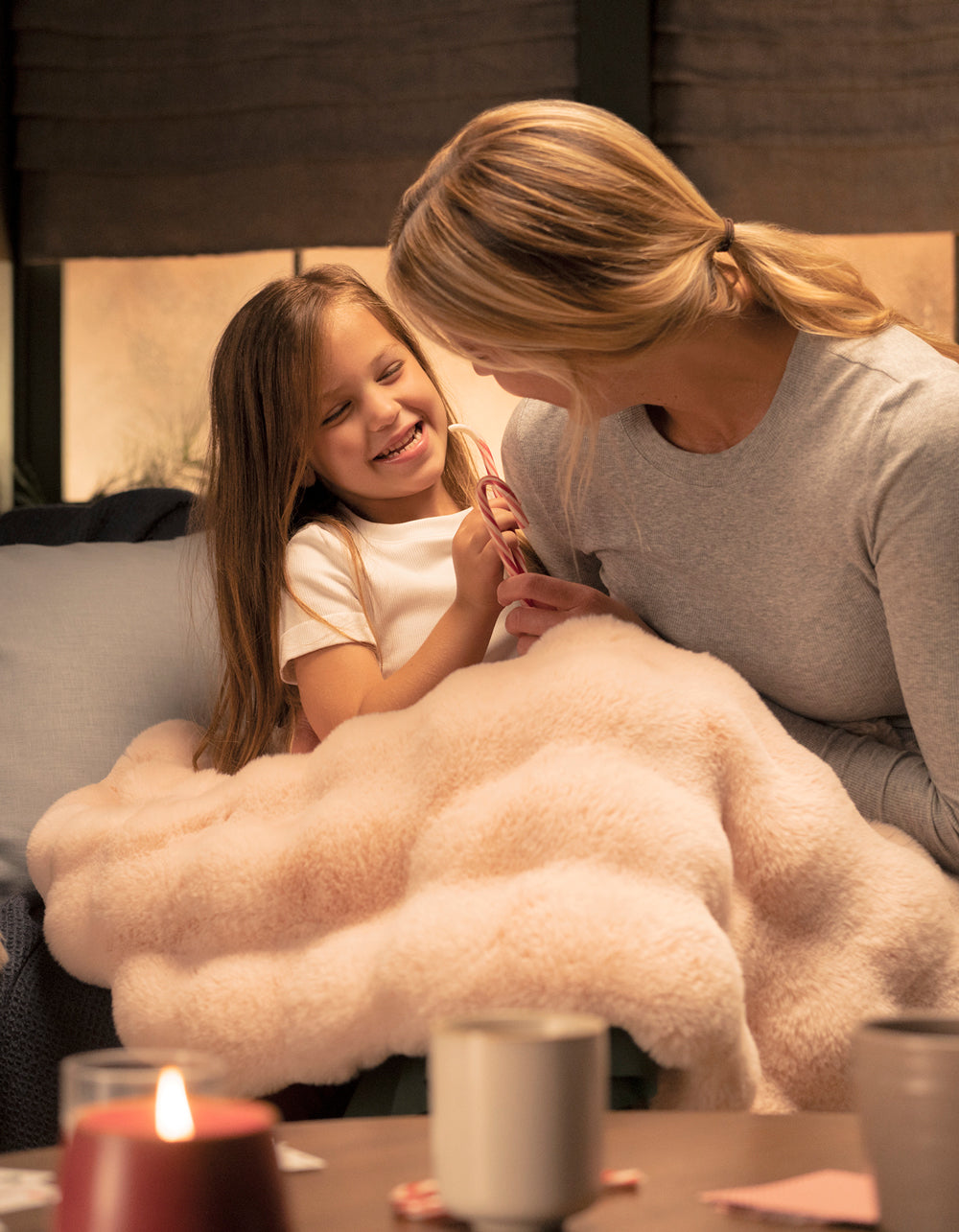 A smiling woman and young girl cuddle under the Cozy Earth Bubble Cuddle Blanket on a couch, sharing a candy cane. Warm lighting and a candle create a relaxed, festive atmosphere.