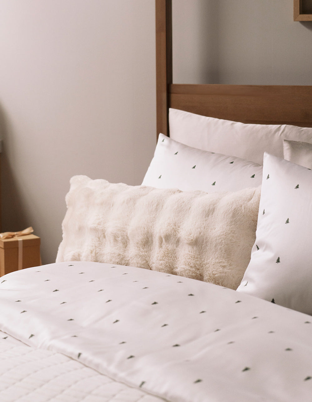 A neatly made bed with white sheets and tree-patterned pillowcases, accented by the Cozy Earth Bubble Cuddle Pillow in cream, set against a wooden headboard in a softly lit room.