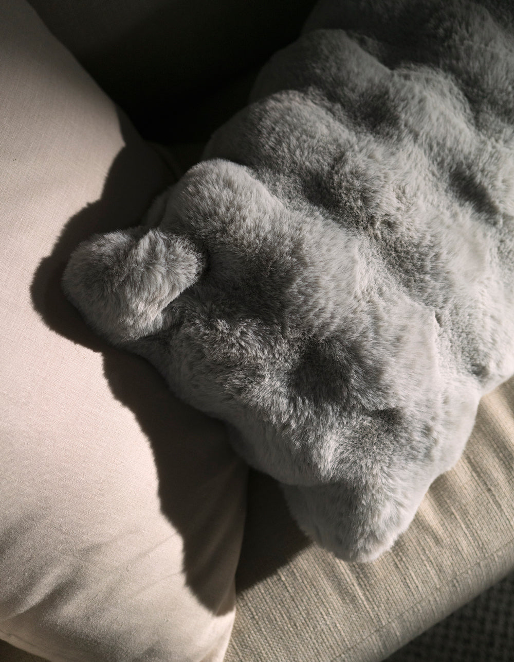The Cozy Earth Bubble Cuddle Pillow, in soft gray faux fur, rests on a beige cushion as sunlight casts gentle shadows across its textured surface.|Color:Light Grey |Size:14" x 30"
