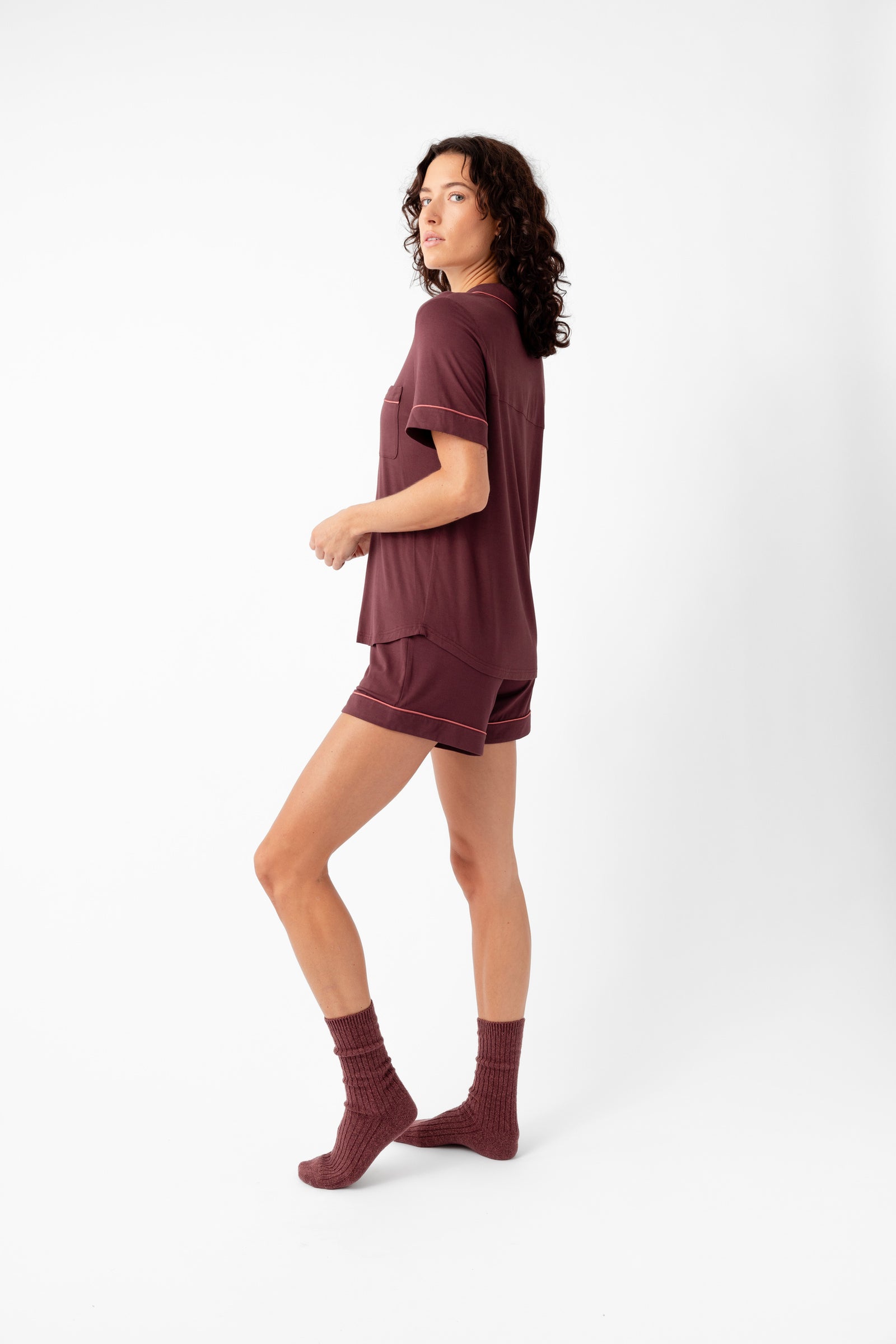 A woman with curly hair stands sideways in a Cozy Earth Women's Bamboo Stretch-Knit Short Sleeve Pajama Set—maroon shorts, short-sleeve top, and socks—against a white background.
